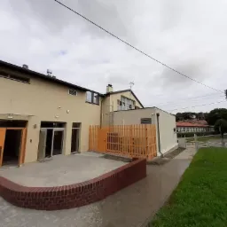 Nowo wybudowany budynek z beżową elewacją, pomarańczowymi akcentami stolarki i ogrodzenia, widok z zewnątrz, w tle pochmurne niebo.