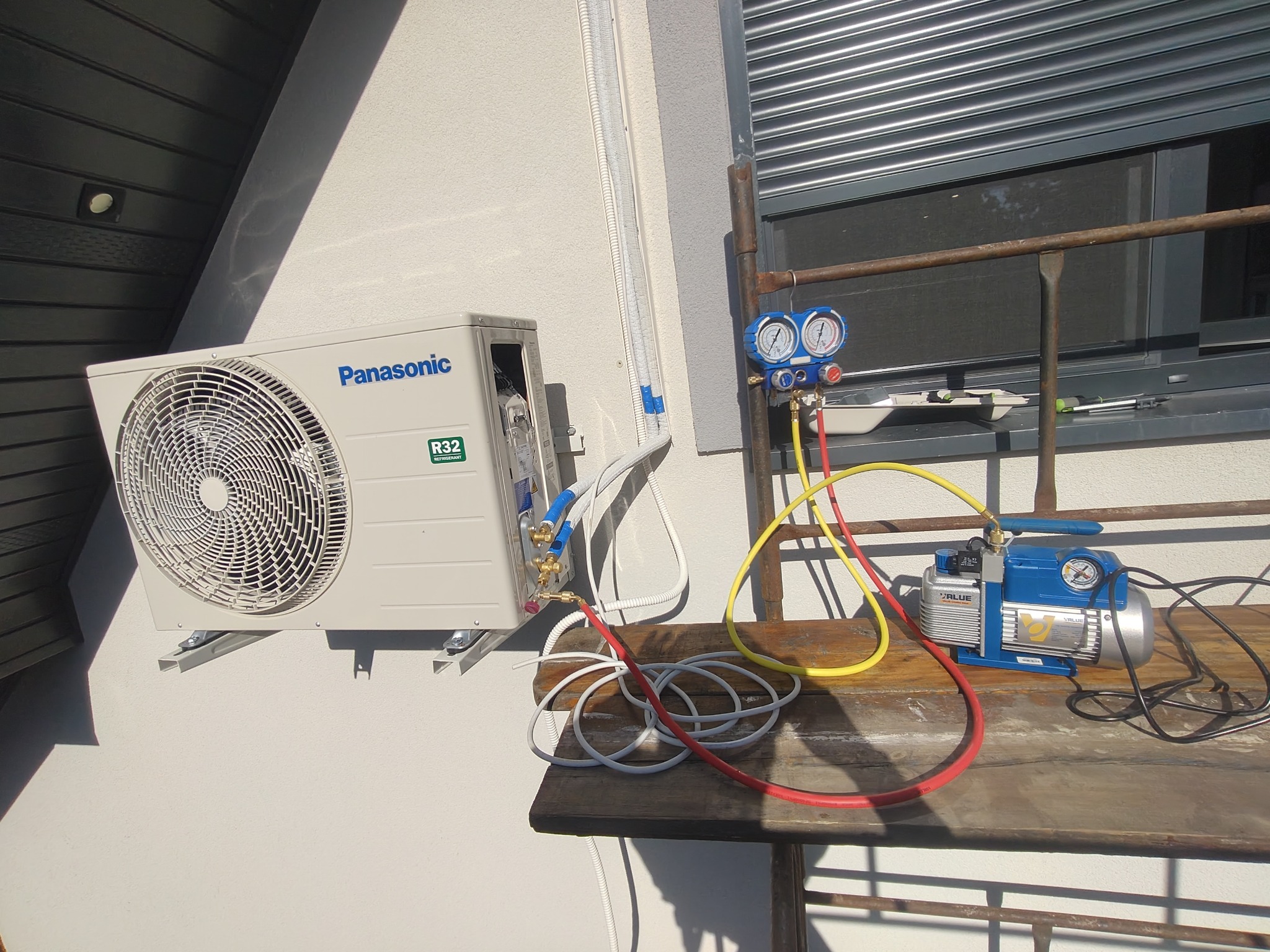Zewnętrzna jednostka klimatyzacji Panasonic R32 zamontowana na ścianie budynku, podłączona do pompy próżniowej i manometru na drewnianym stole podczas serwisu.