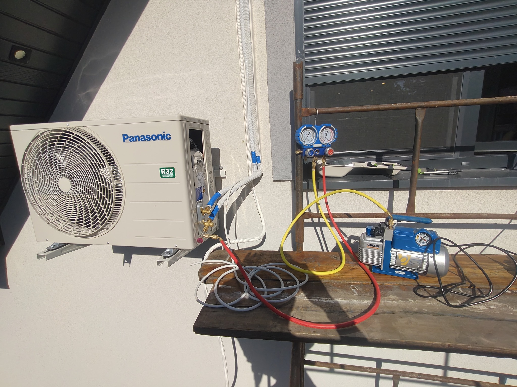 Biały klimatyzator Panasonic R32 zamontowany na ścianie, podłączony do pompy próżniowej i manometrów na zewnątrz budynku. Widoczne przewody i narzędzia serwisowe.