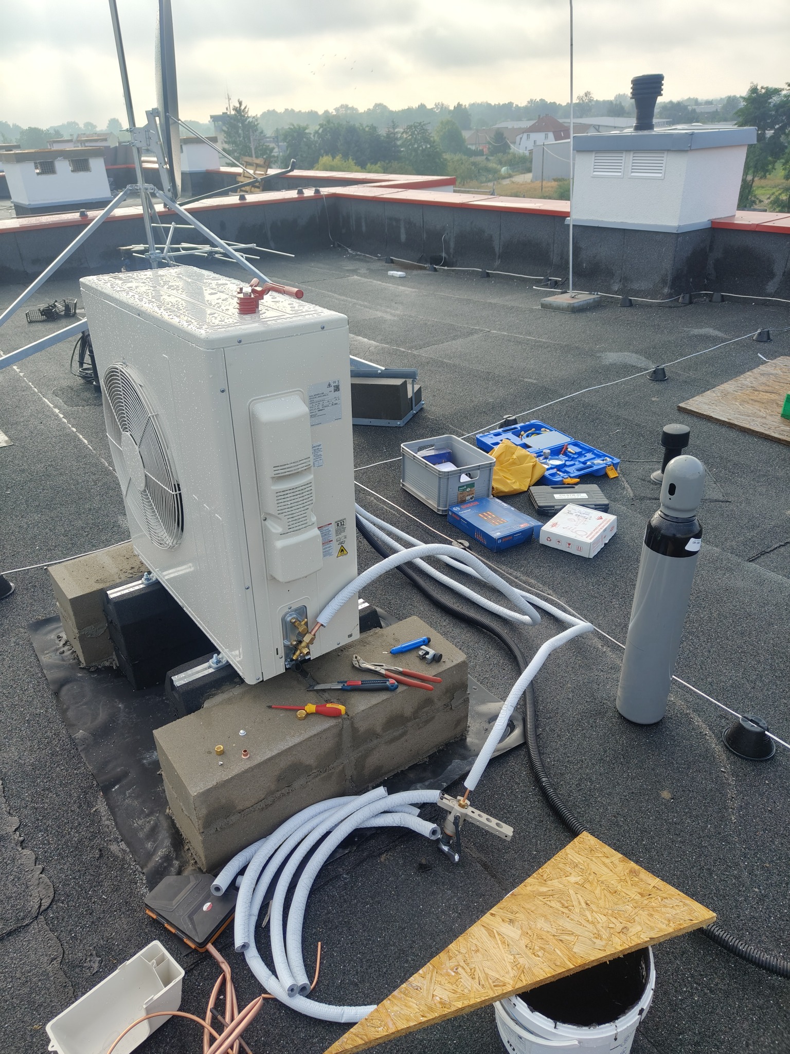 Biała jednostka zewnętrzna klimatyzacji na dachu, w trakcie montażu. Widoczne narzędzia, rury, butla z gazem i elementy montażowe. Dach pokryty papą.