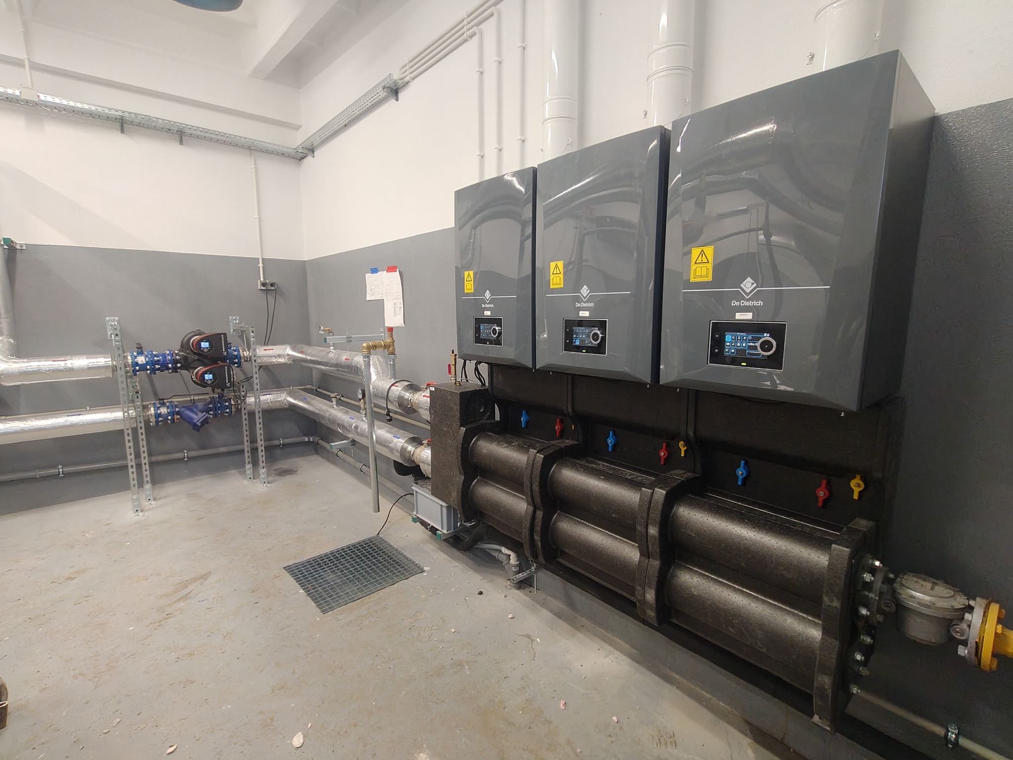Kompletna instalacja grzewcza z trzema szarymi piecami De Dietrich, rurami w izolacji i zaworami. Widoczne elementy instalacji hydraulicznej oraz odpływy wentylacyjne.