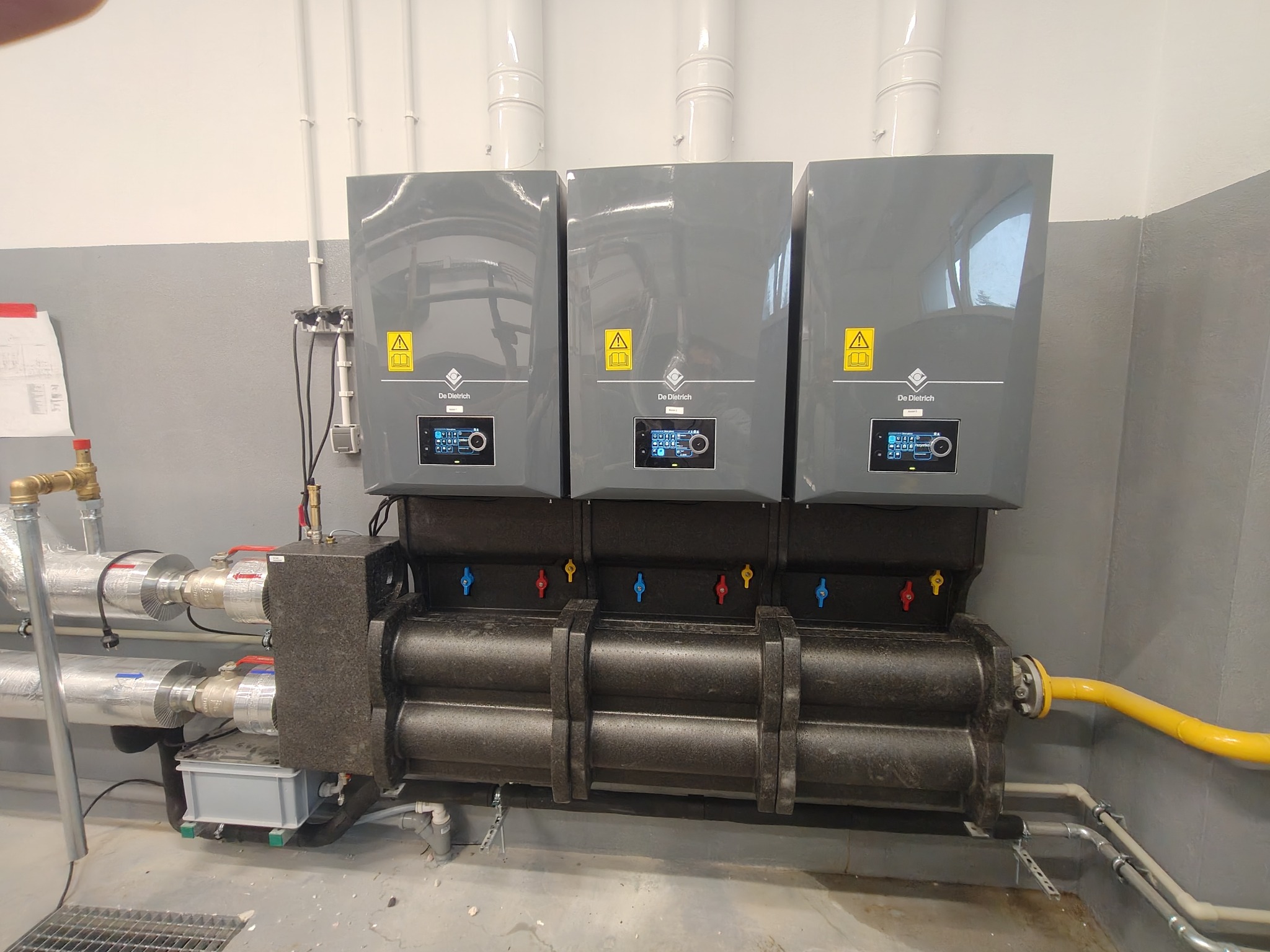 Trzy szare, wiszące piece gazowe De Dietrich z podłączonymi rurami, zaworami i przewodami, zainstalowane w szarej kotłowni. Widoczne detale instalacji.