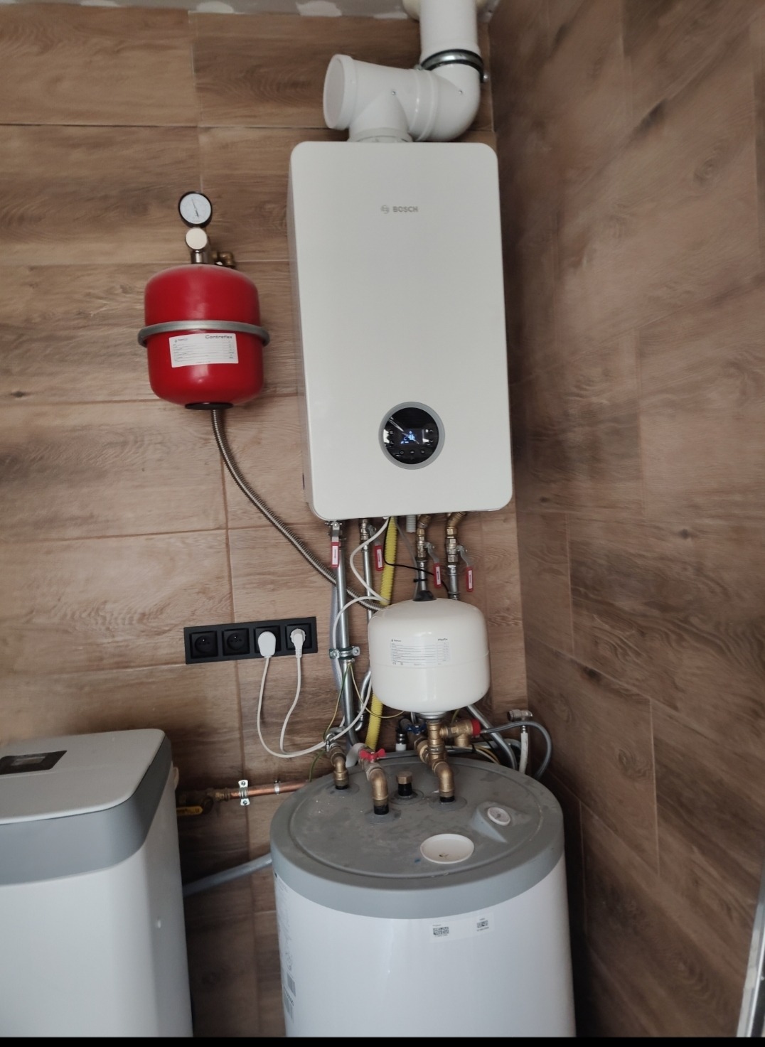 Zamontowany, nowoczesny, biały piec gazowy marki Bosch z czerwonym zbiornikiem wyrównawczym na ścianie z drewnopodobnych płytek. Widoczne podłączenia hydrauliczne i elektryczne.