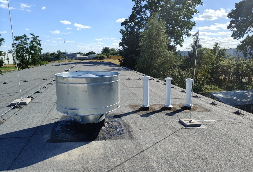 Metalowy komin wentylacyjny na płaskim dachu pokrytym papą, obok trzy białe rury wentylacyjne. Na dachu widoczne elementy odgromowe i antena.