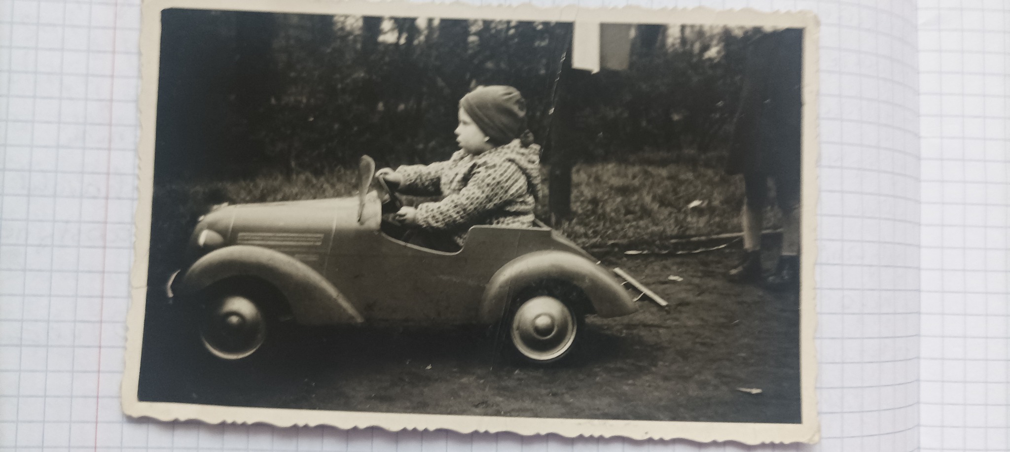 Czarno-biała fotografia dziecka w zabawkowej replice samochodu w Biłgoraju. Ujęcie z profilu, widoczne detale pojazdu i ubrania dziecka. Tło stanowią drzewa i trawa.