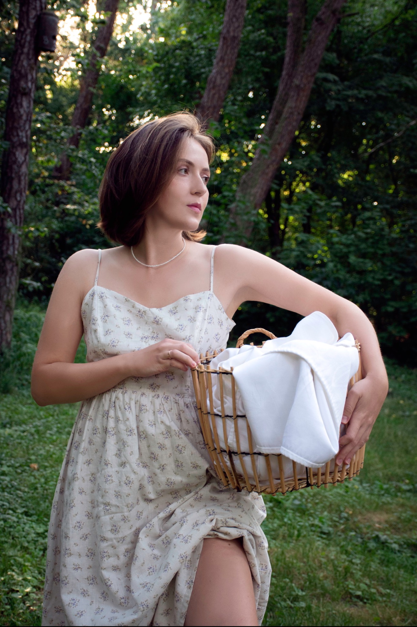 Kobieta w kwiecistej sukience, trzyma wiklinowy kosz z białymi tkaninami, stojąc na tle zieleni drzew i promieni słonecznych.