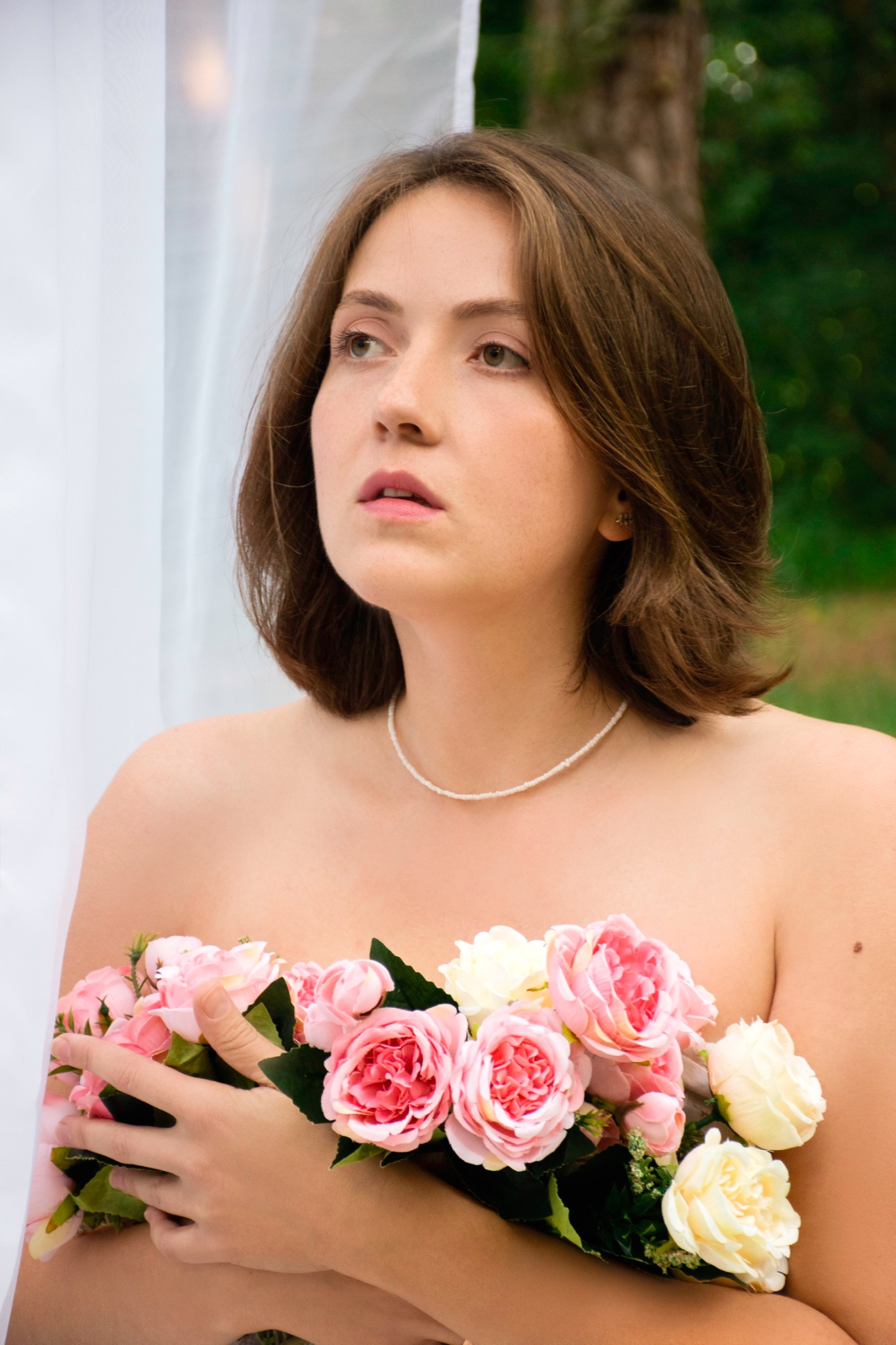 Portret kobiety z bukietem różowych i białych róż, noszącej delikatny naszyjnik z koralików, na tle białej zasłony i zieleni.