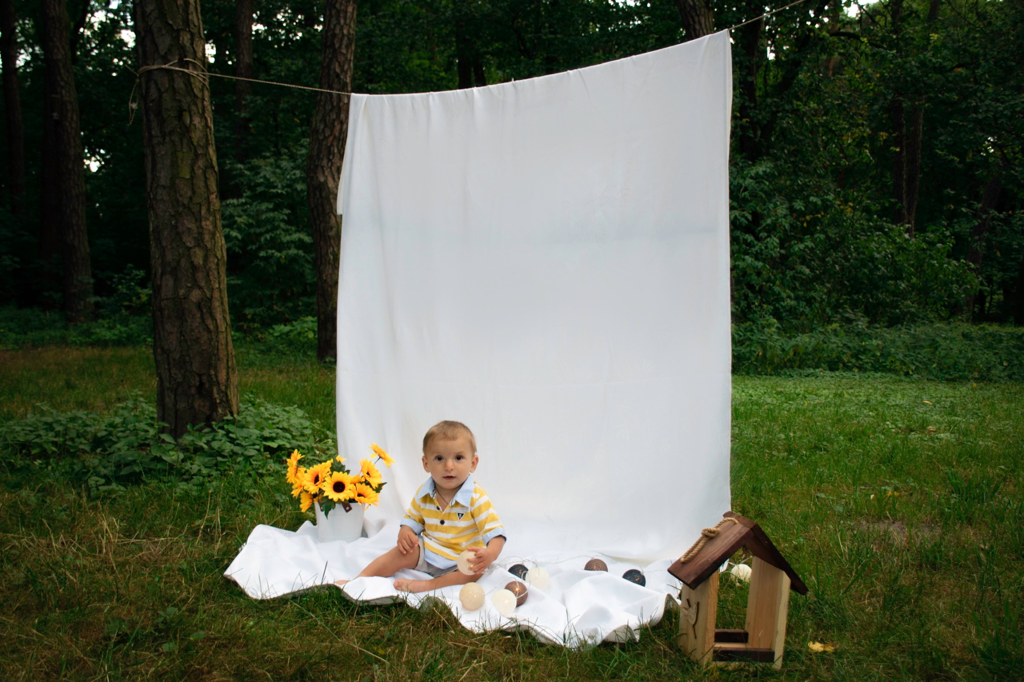 Chłopiec w pasiastym polo siedzi na białym płótnie w plenerze, obok wiadro słoneczników i drewniany karmnik dla ptaków, białe tło rozwieszone na drzewach.