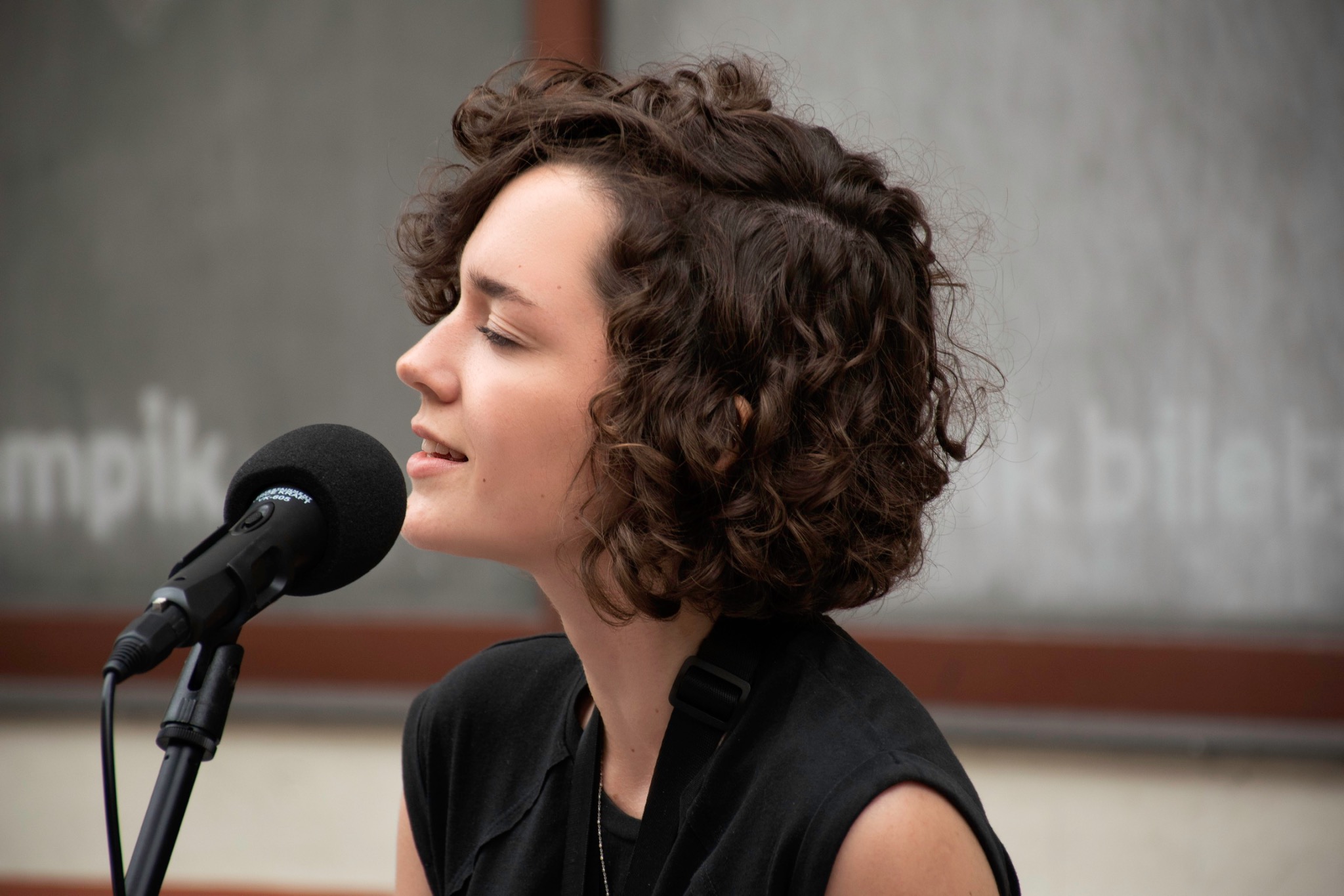 Profil kobiety z kręconymi włosami śpiewającej do mikrofonu na statywie, z widocznym napisem 'mpik' w tle.