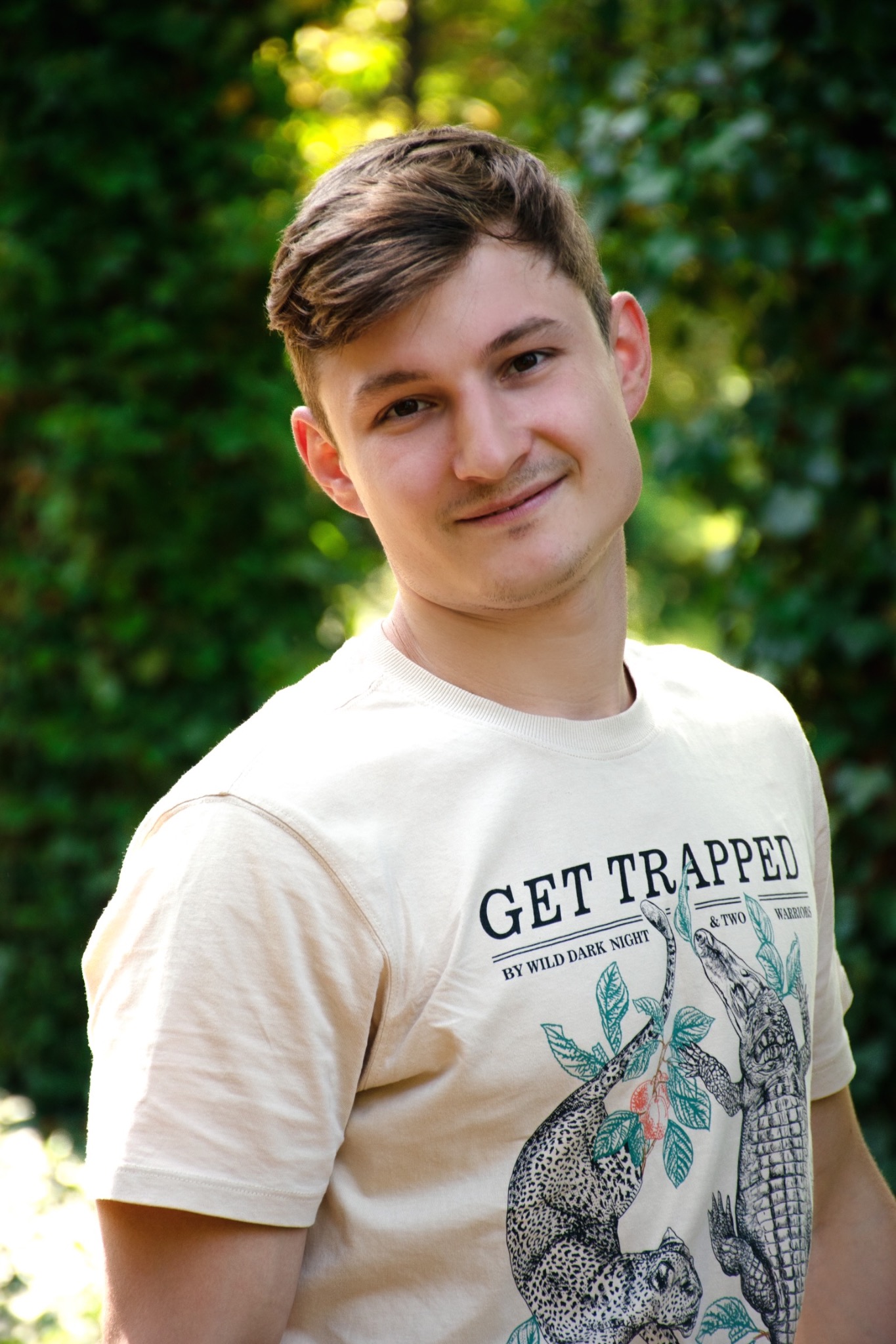 Portret młodego mężczyzny w beżowej koszulce z nadrukiem przedstawiającym lamparta i krokodyla na tle zieleni.