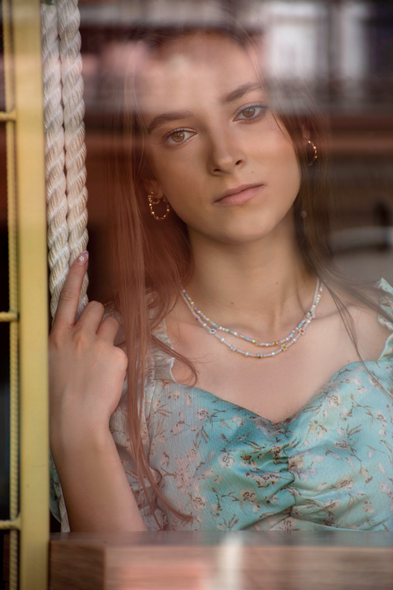 Portret młodej kobiety z delikatnym makijażem, w kwiecistej sukience i z dwoma naszyjnikami z koralików, stojącej za oknem z żółtą ramą i grubym sznurem. Na paznokciach subtelny manicure...