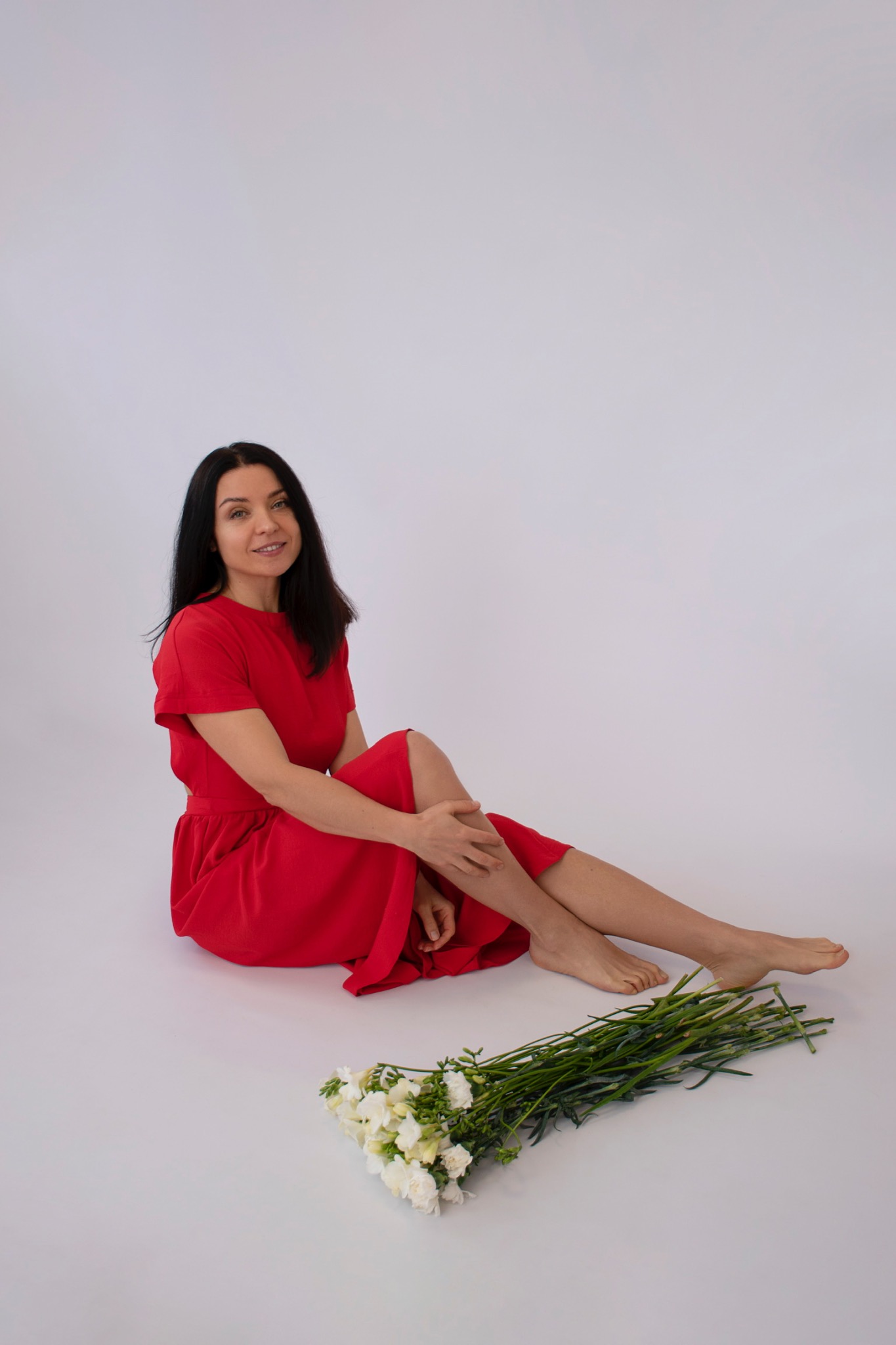 Kobieta w czerwonej sukience siedzi na podłodze z bukietem białych frezji na jasnym tle.