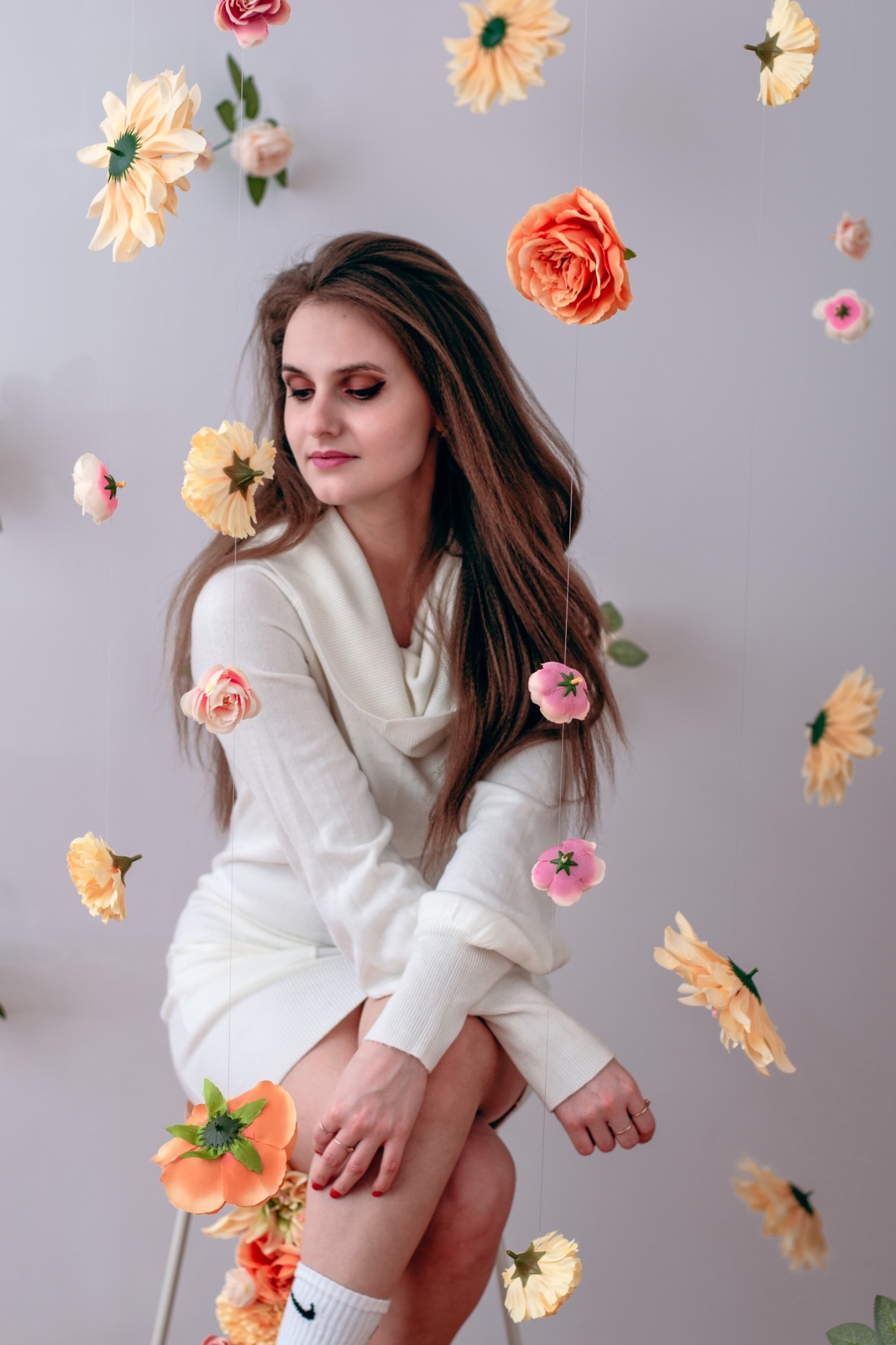 Portret kobiety w białej sukience, otoczonej wiszącymi sztucznymi kwiatami w odcieniach różu i pomarańczy, na jasnym tle.
