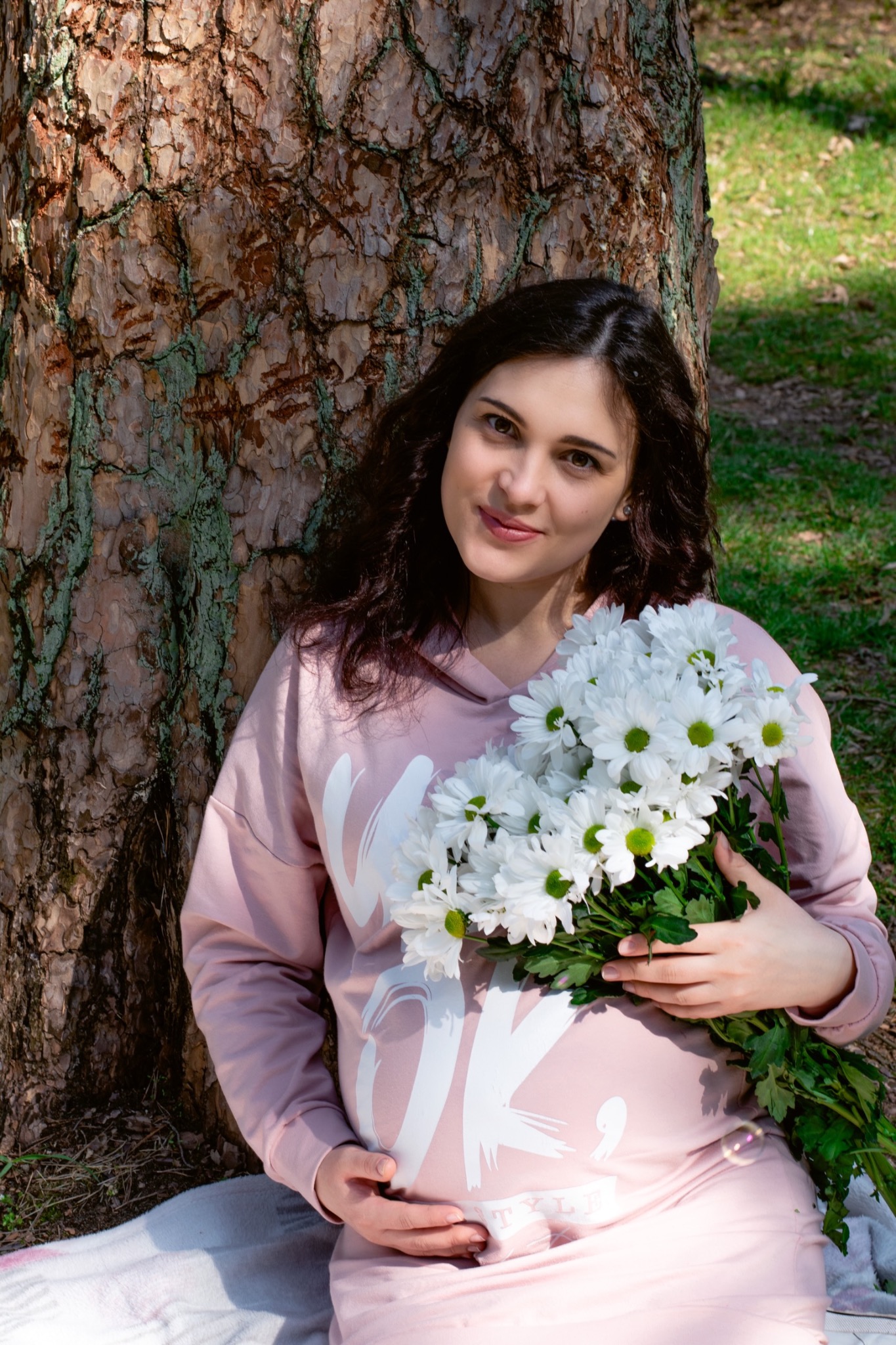 Uśmiechnięta, ciężarna kobieta w różowej bluzie, opiera się o drzewo i trzyma bukiet białych chryzantem.