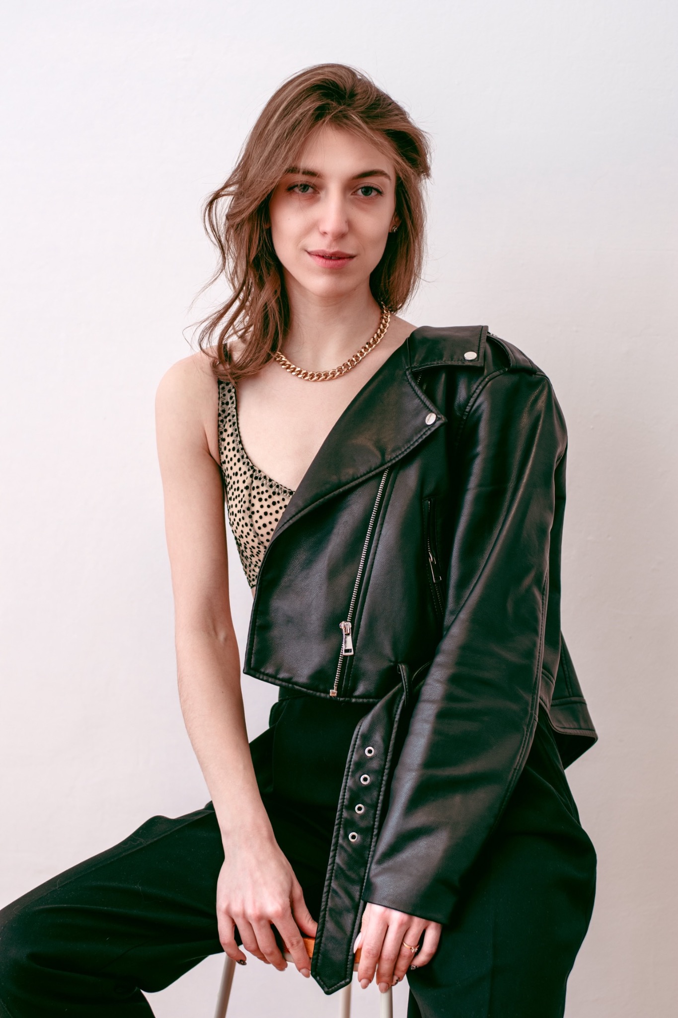 Portret kobiety siedzącej na krześle, ubrana w top w kropki, czarne spodnie i skórzaną kurtkę nonszalancko zarzuconą na ramię, z grubym złotym łańcuchem na szyi.
