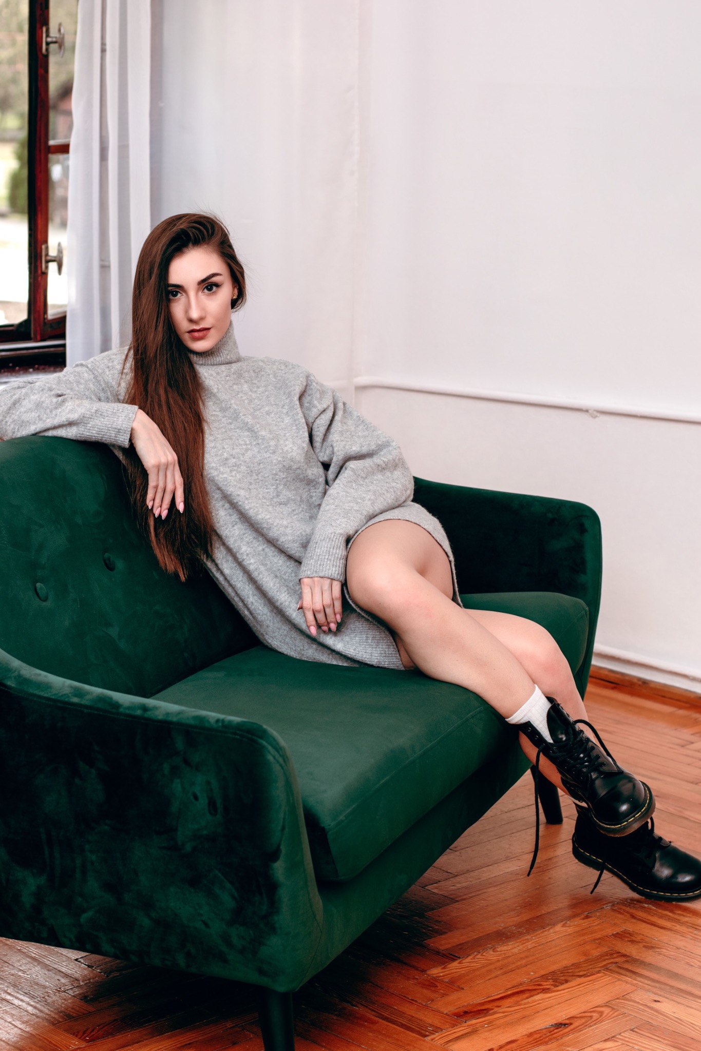 Kobieta w szarym swetrze i czarnych butach siedzi na zielonym, welurowym fotelu w jasnym wnętrzu.