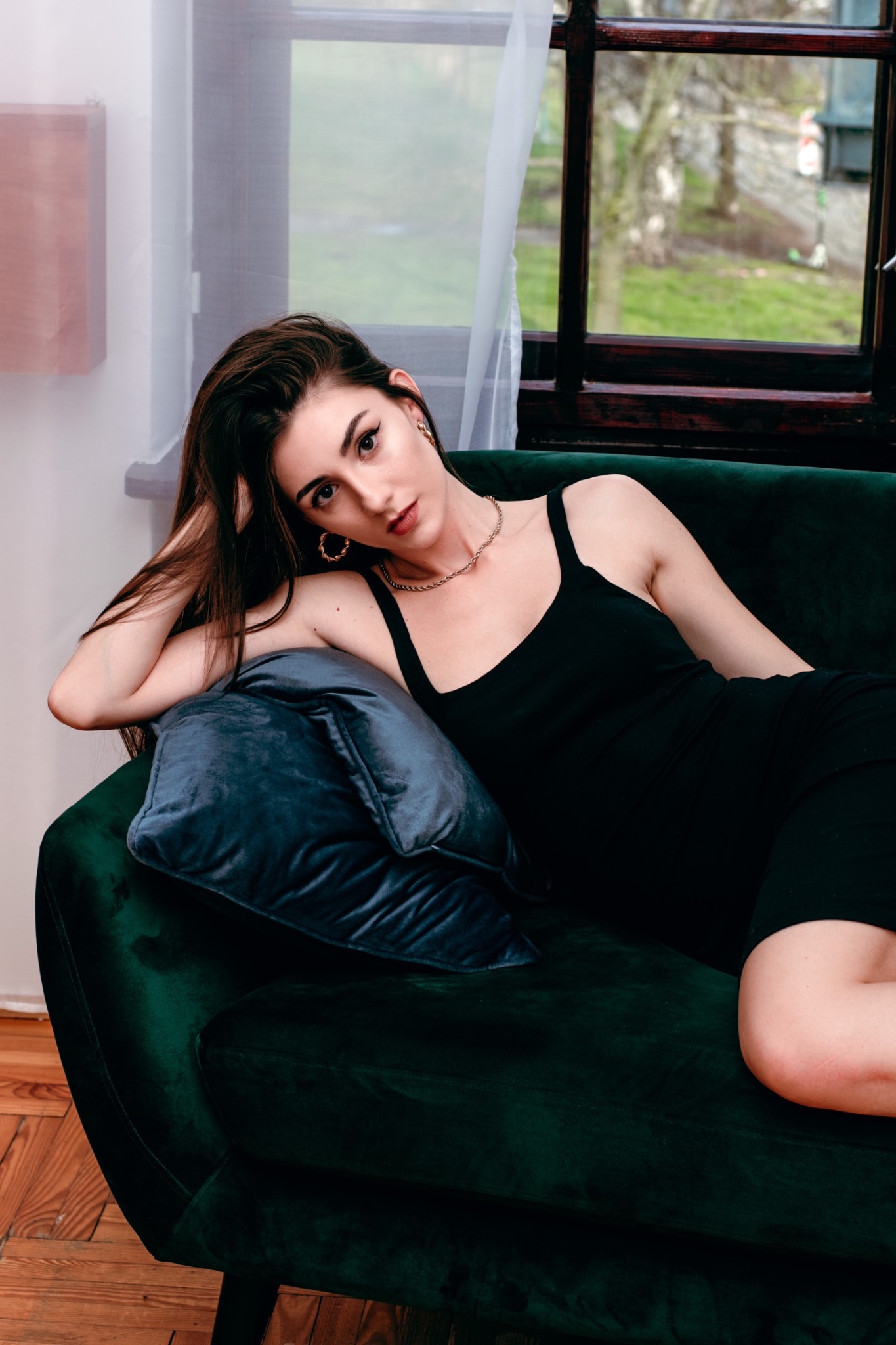 Kobieta w czarnej sukience leży na zielonej, aksamitnej sofie z poduszką na tle okna z widokiem na zieleń.