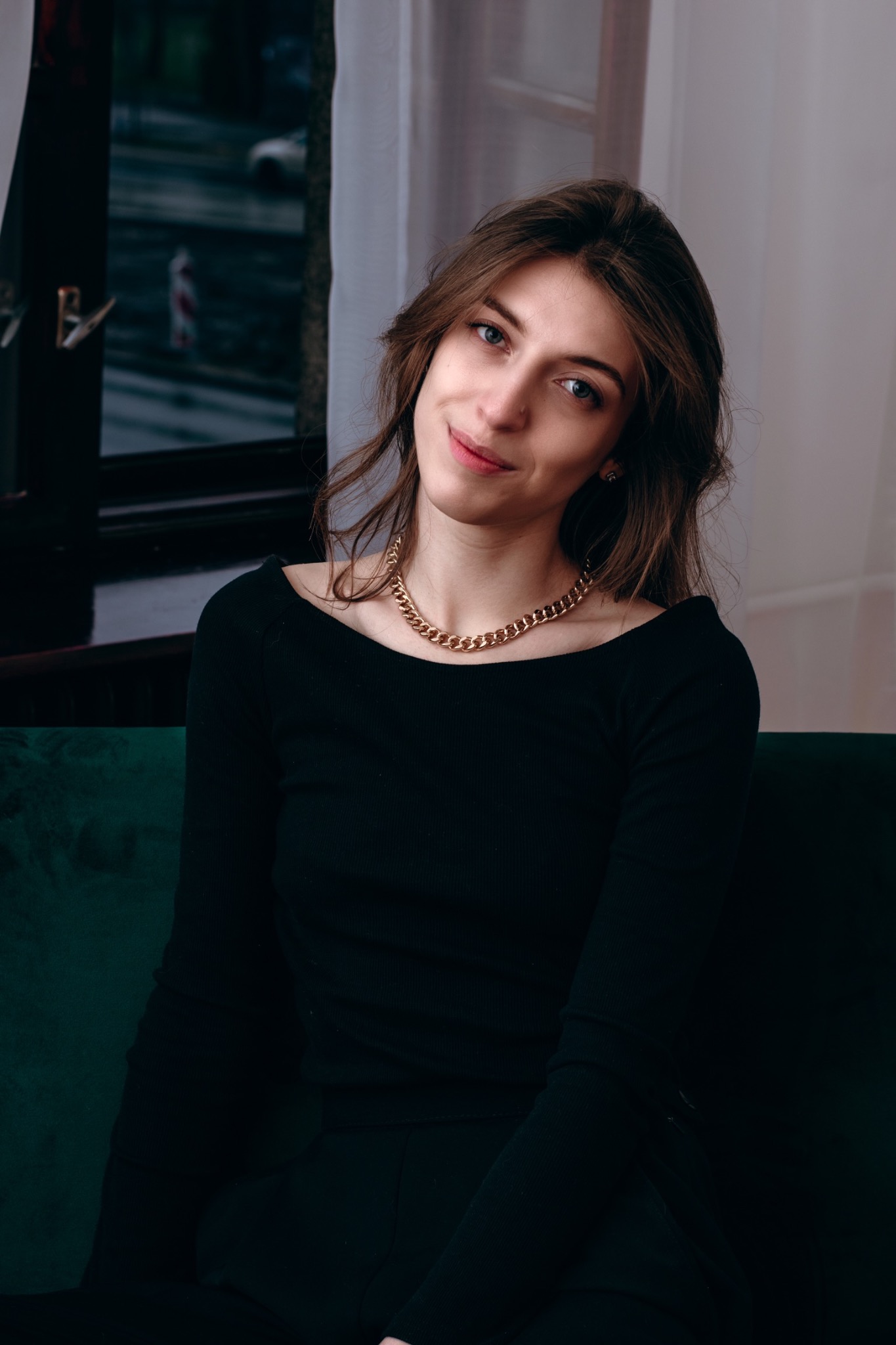 Portret kobiety z delikatnym uśmiechem, w czarnej bluzce z odkrytymi ramionami i grubym złotym łańcuchem, siedzącej na ciemnozielonej sofie na tle okna z białymi zasłonami.