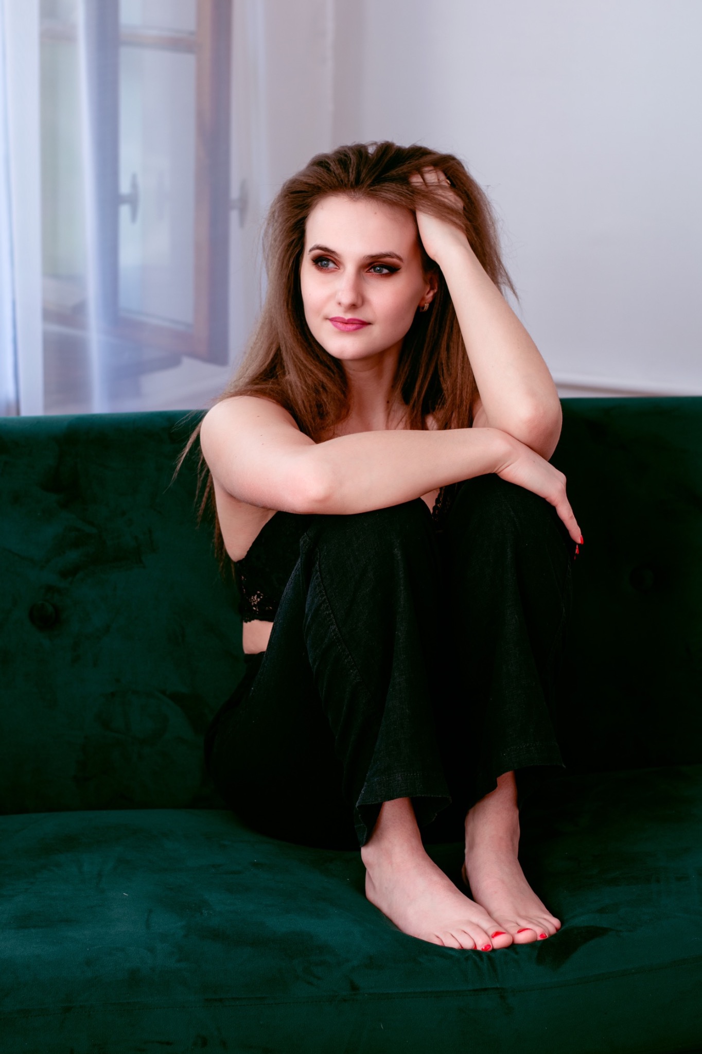 Portret kobiety w czarnym stroju, siedzącej na zielonej welurowej kanapie, z ręką we włosach i zamyślonym spojrzeniem w stronę okna.