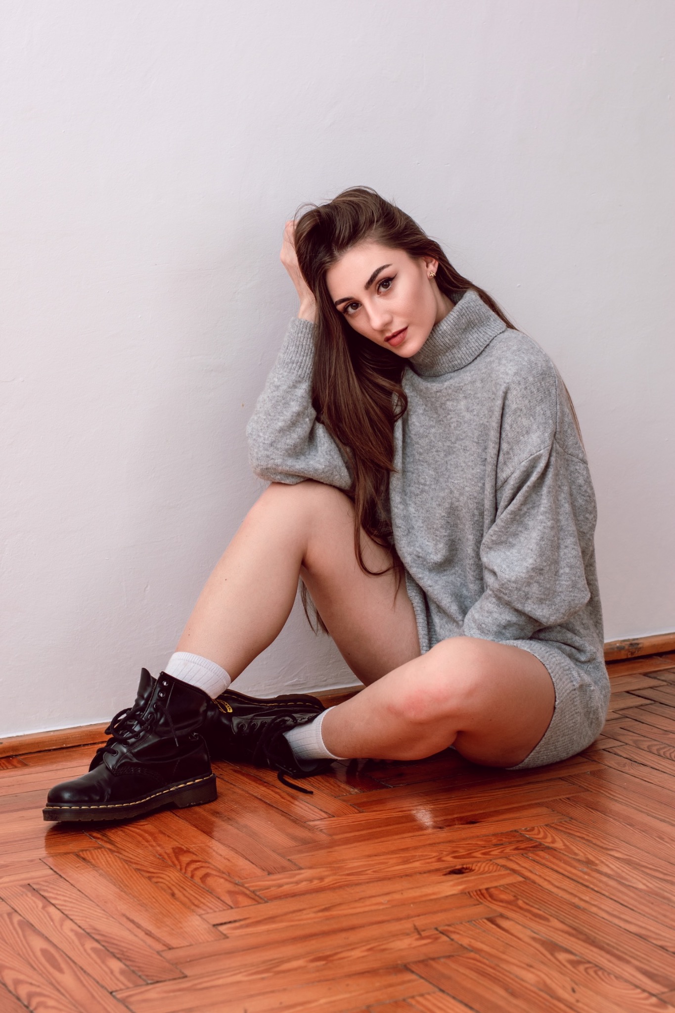 Portret kobiety w szarym swetrze i czarnych butach siedzącej na parkiecie, opierającej głowę o dłoń, z melancholijnym spojrzeniem.