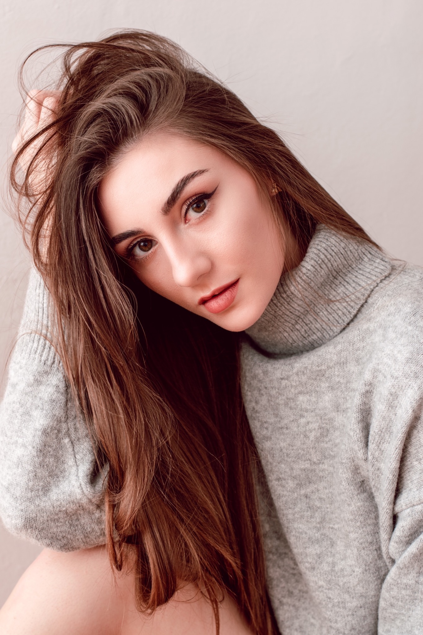 Portret kobiety w szarym swetrze z golfem, z ręką we włosach, patrzącej w dół.