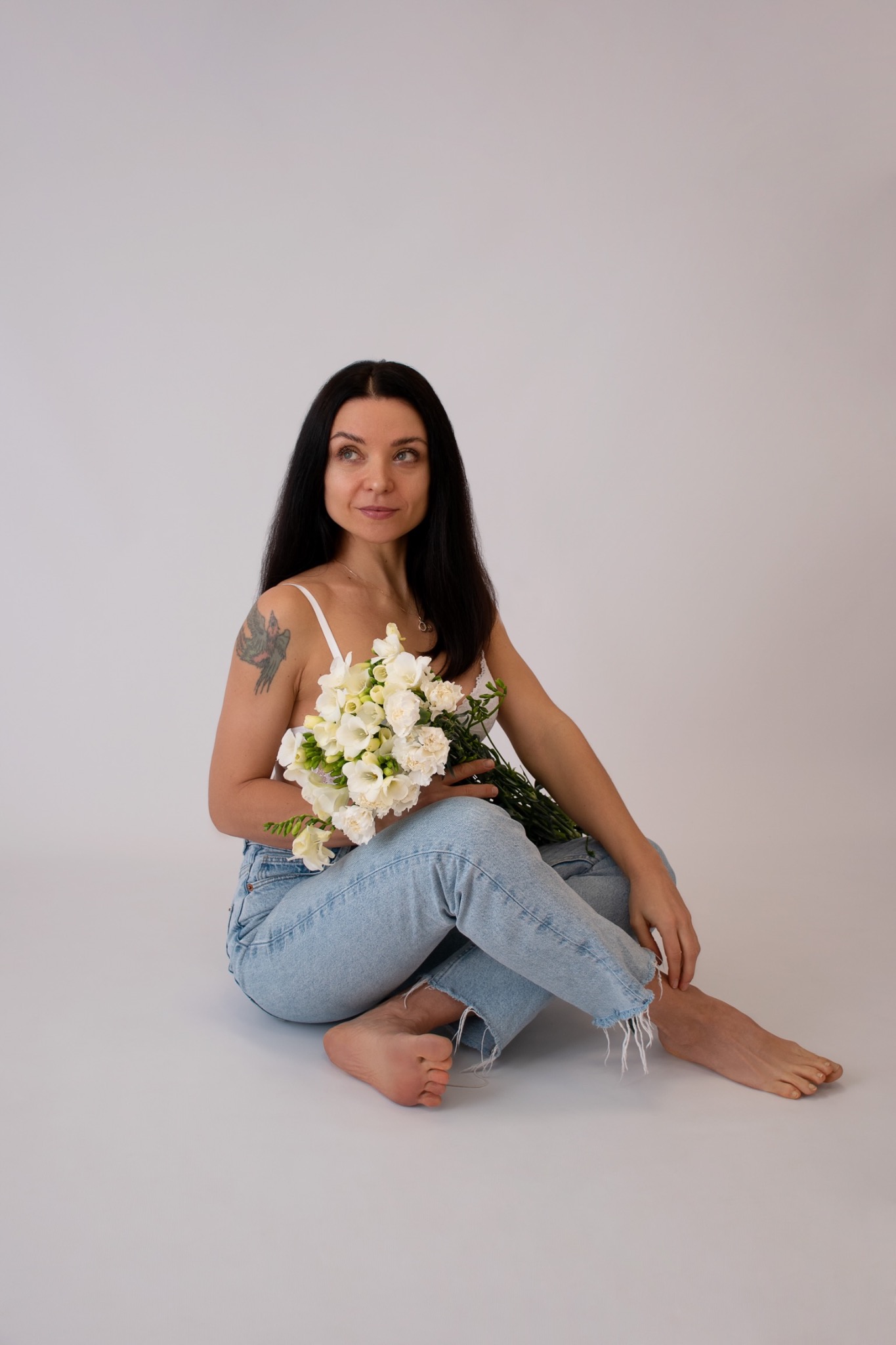 Kobieta siedzi na podłodze na jasnym tle, ubrana w dżinsy i białą bieliznę, trzyma bukiet białych frezji, z tatuażem na ramieniu i delikatnym uśmiechem.