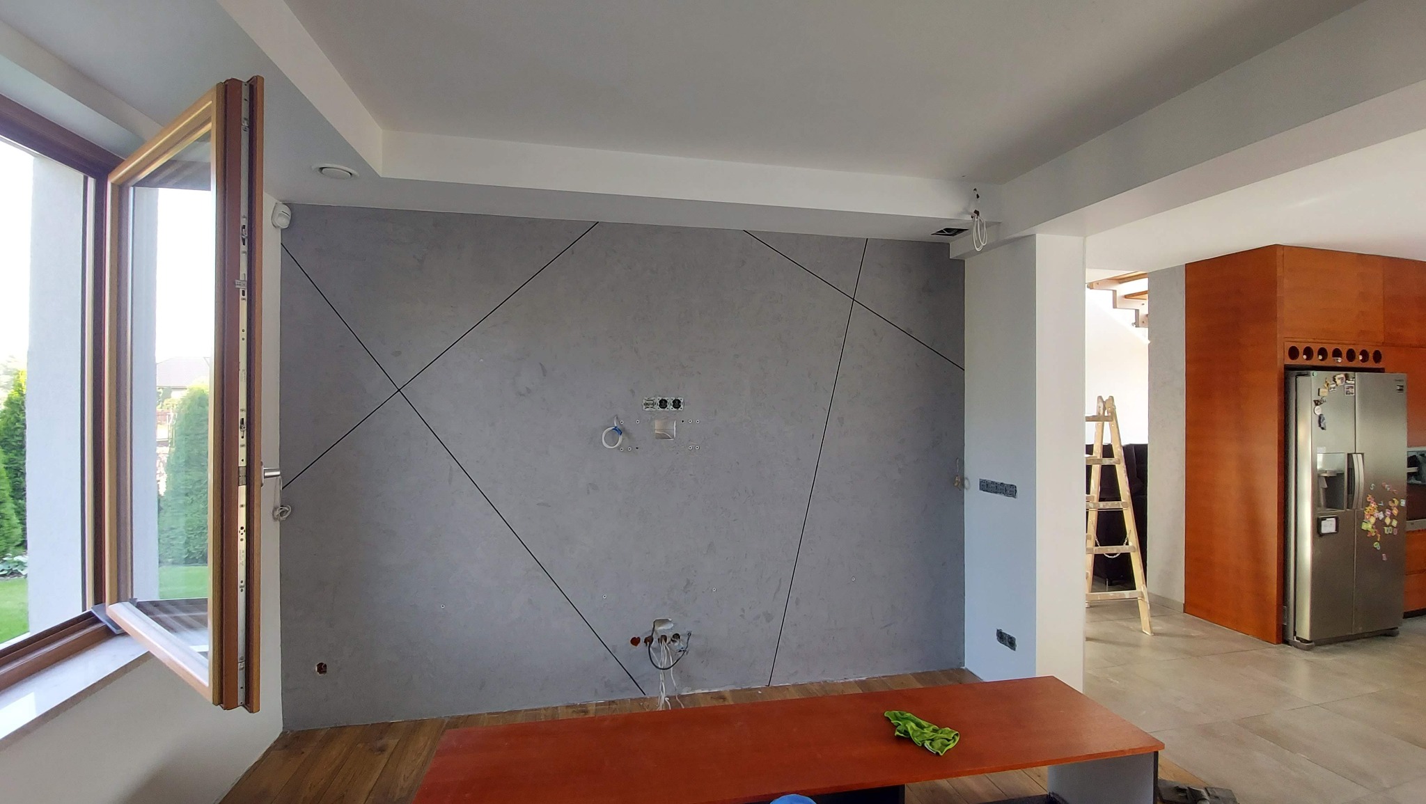 Surowa ściana w szarym odcieniu z geometrycznym wzorem z czarnych linii, widoczne gniazdka elektryczne i wyprowadzenia hydrauliczne, w tle otwarta przestrzeń z drewnianymi elementami i drabina.