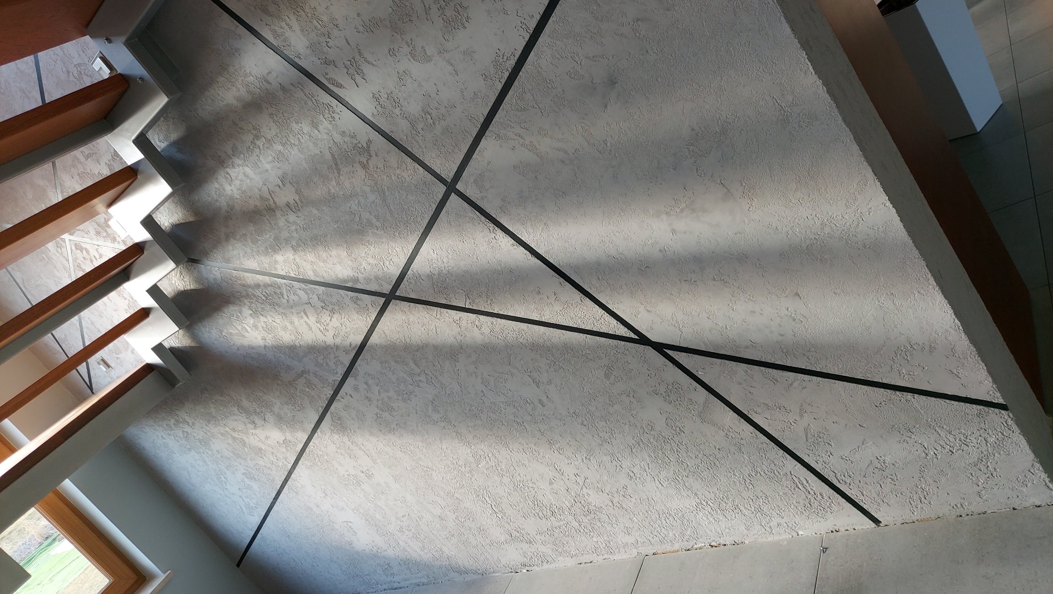 Dekoracyjna ściana o fakturze betonu z czarnymi, ukośnymi listwami, widok z dołu, fragment schodów z drewnianymi stopniami po lewej stronie.