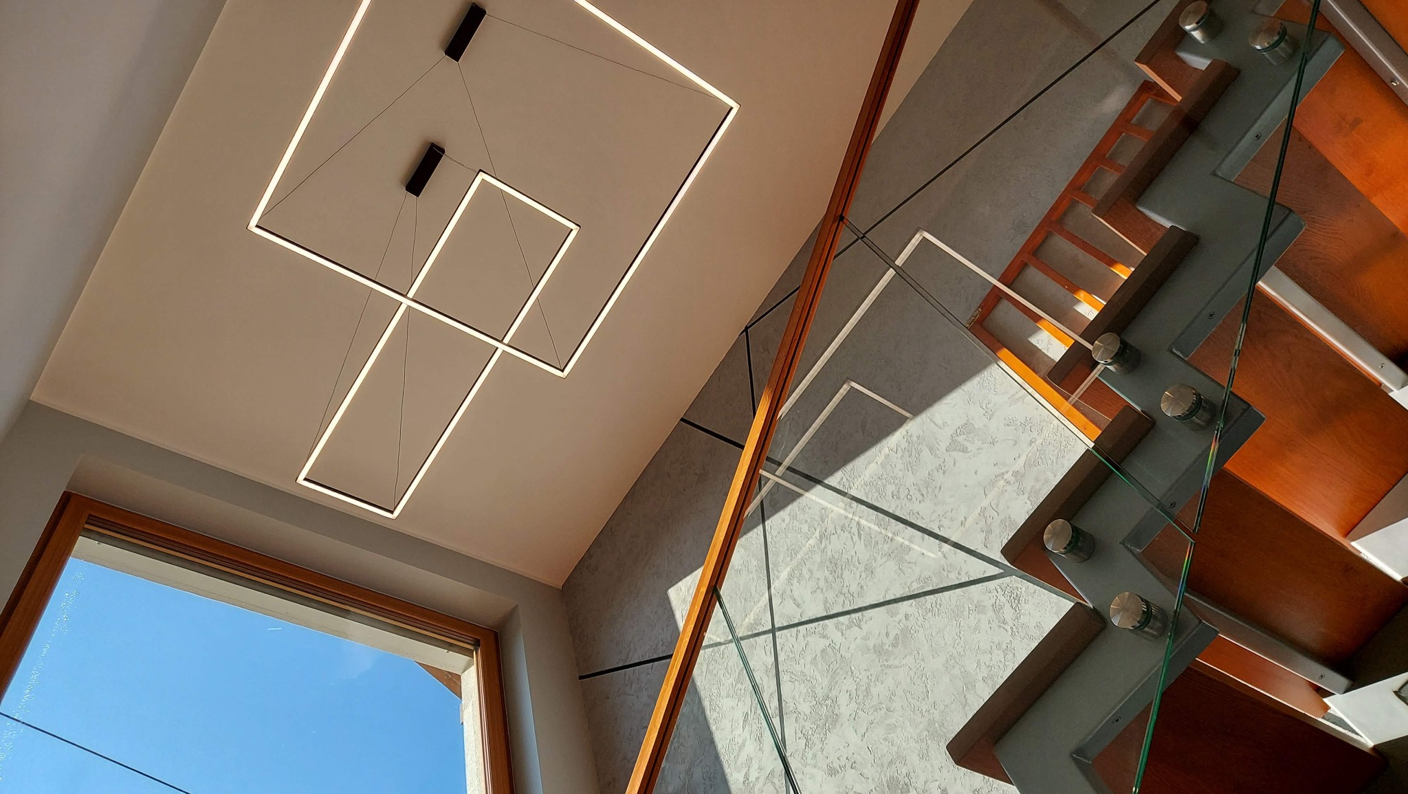 Nowoczesne schody drewniane z balustradą ze szkła, oświetlenie sufitowe w kształcie geometrycznych kwadratów, widok z dołu na błękitne niebo przez okno.