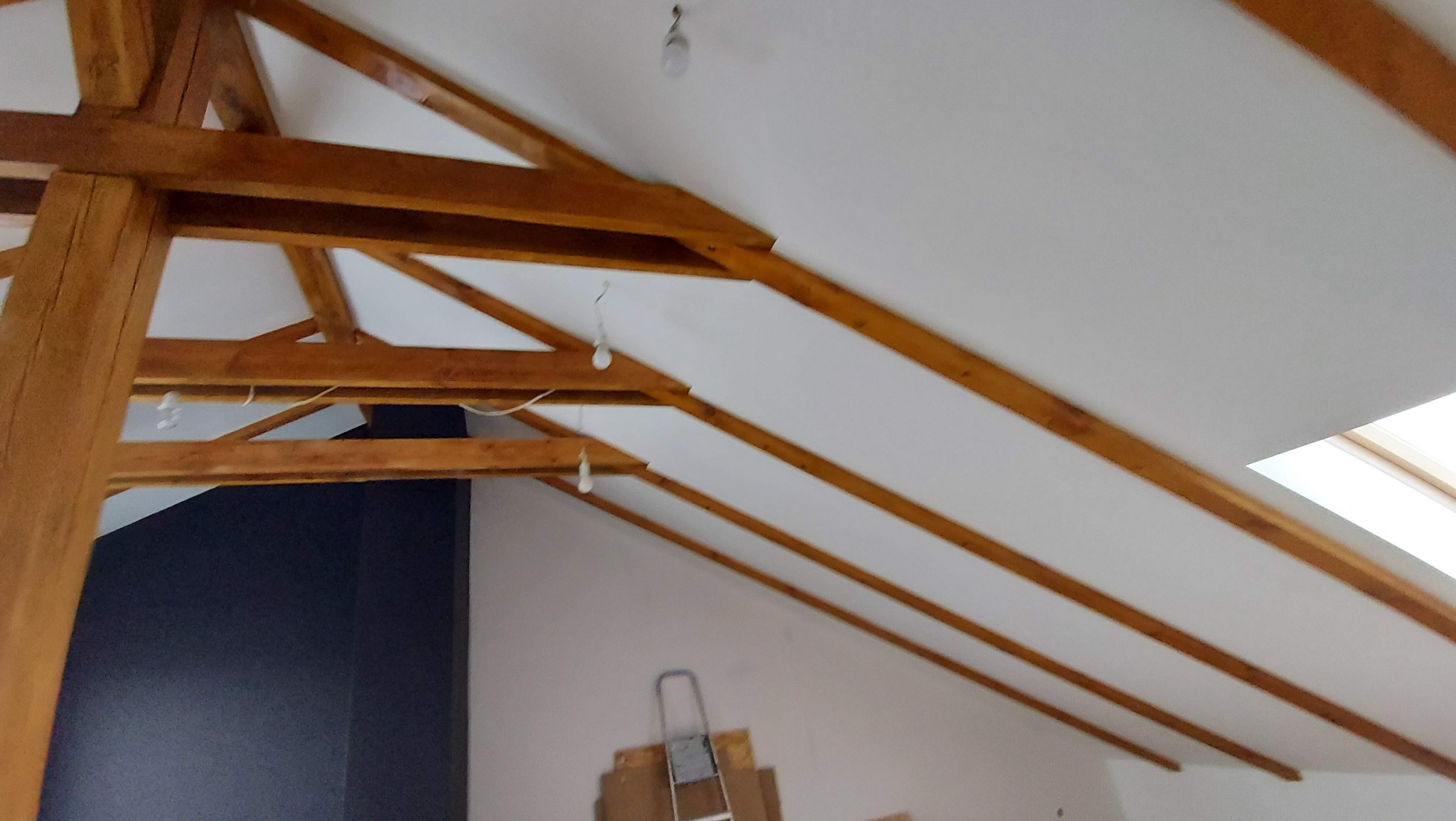 Skośny sufit poddasza z drewnianymi belkami konstrukcyjnymi, częściowo pomalowany na biało, z widocznym oknem dachowym i wiszącymi żarówkami, w tle ściana pomalowana na ciemny kolor.