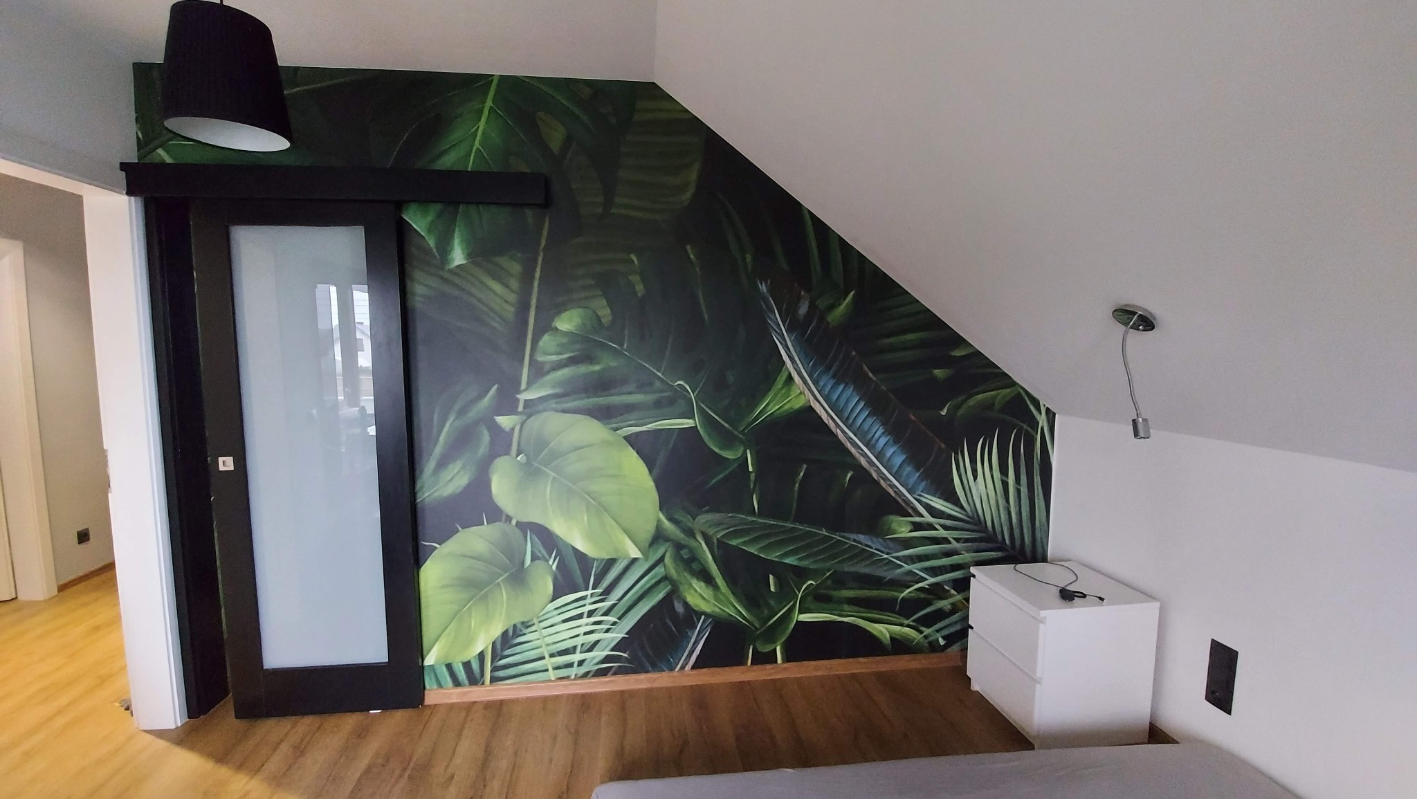 Pokój w Poznaniu z fototapetą w motywy roślinne na ścianie, przesuwne drzwi z czarną ramą i biała szafka nocna pod skosem dachu.