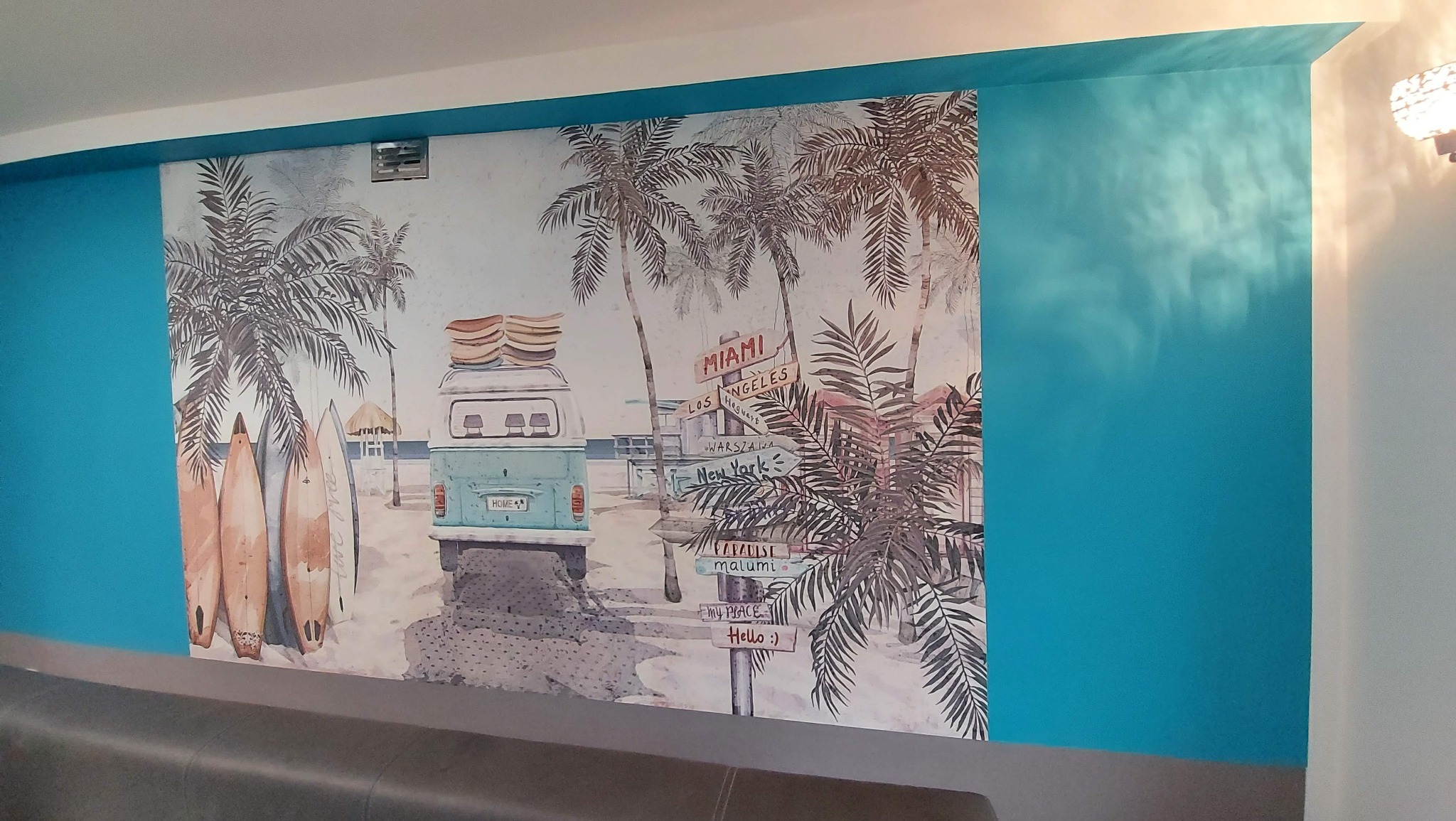 Malowidło ścienne przedstawiające plażowy motyw z palmami, surferami, turkusowym vanem z deskami surfingowymi na dachu, oraz drogowskazami do różnych miast świata, w tym Miami, Los Angeles...