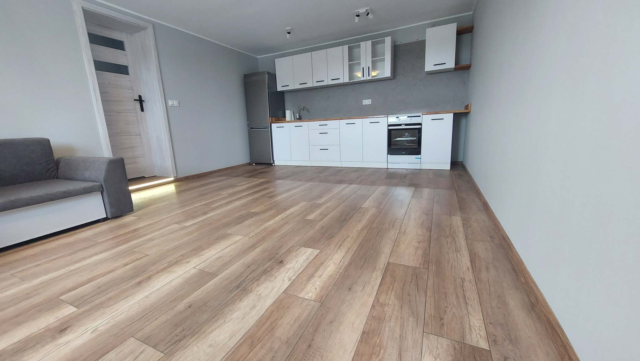 Widok ogólny nowocześnie urządzonej kuchni z drewnopodobną podłogą, białymi szafkami i szarą ścianą, z fragmentem sofy w rogu pokoju i widocznymi drzwiami po lewej stronie.