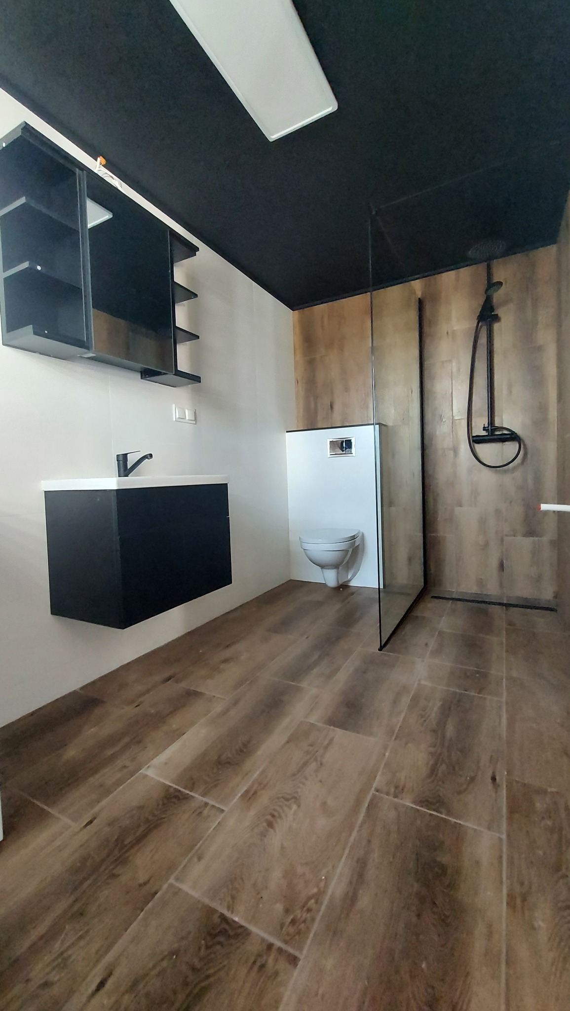 Nowoczesna łazienka z czarnym sufitem, drewnopodobną podłogą i szklaną kabiną prysznicową typu walk-in, wyposażona w wiszącą szafkę z umywalką i toaletę podtynkową.