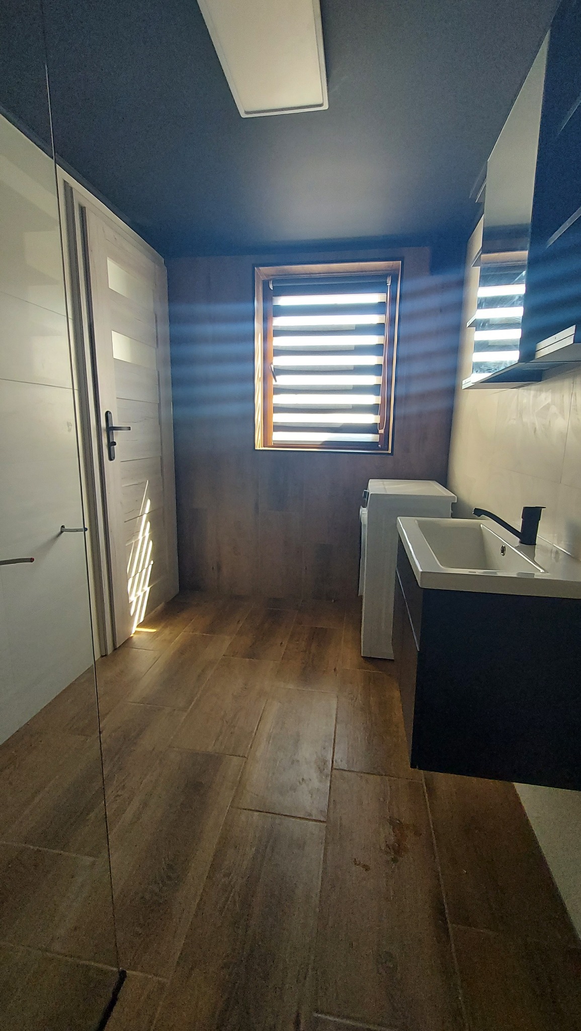 Nowoczesna łazienka z drewnopodobną podłogą, ciemnoszarym sufitem i oknem z żaluzjami, wyposażona w białą pralkę, umywalkę z szafką i kabinę prysznicową.