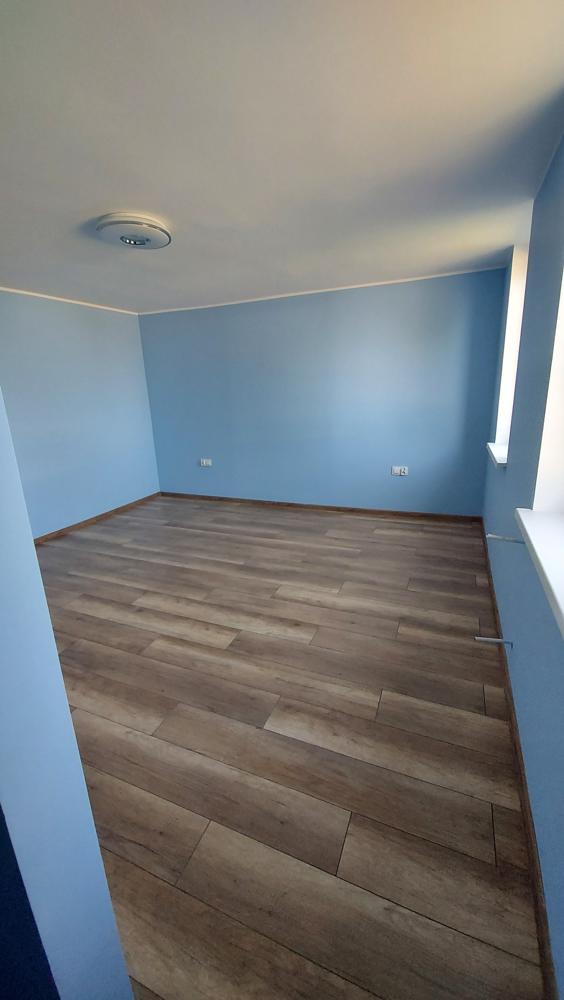 Pusty pokój z błękitnymi ścianami i podłogą z paneli imitujących drewno, widoczne gniazdka elektryczne i minimalistyczna lampa sufitowa.