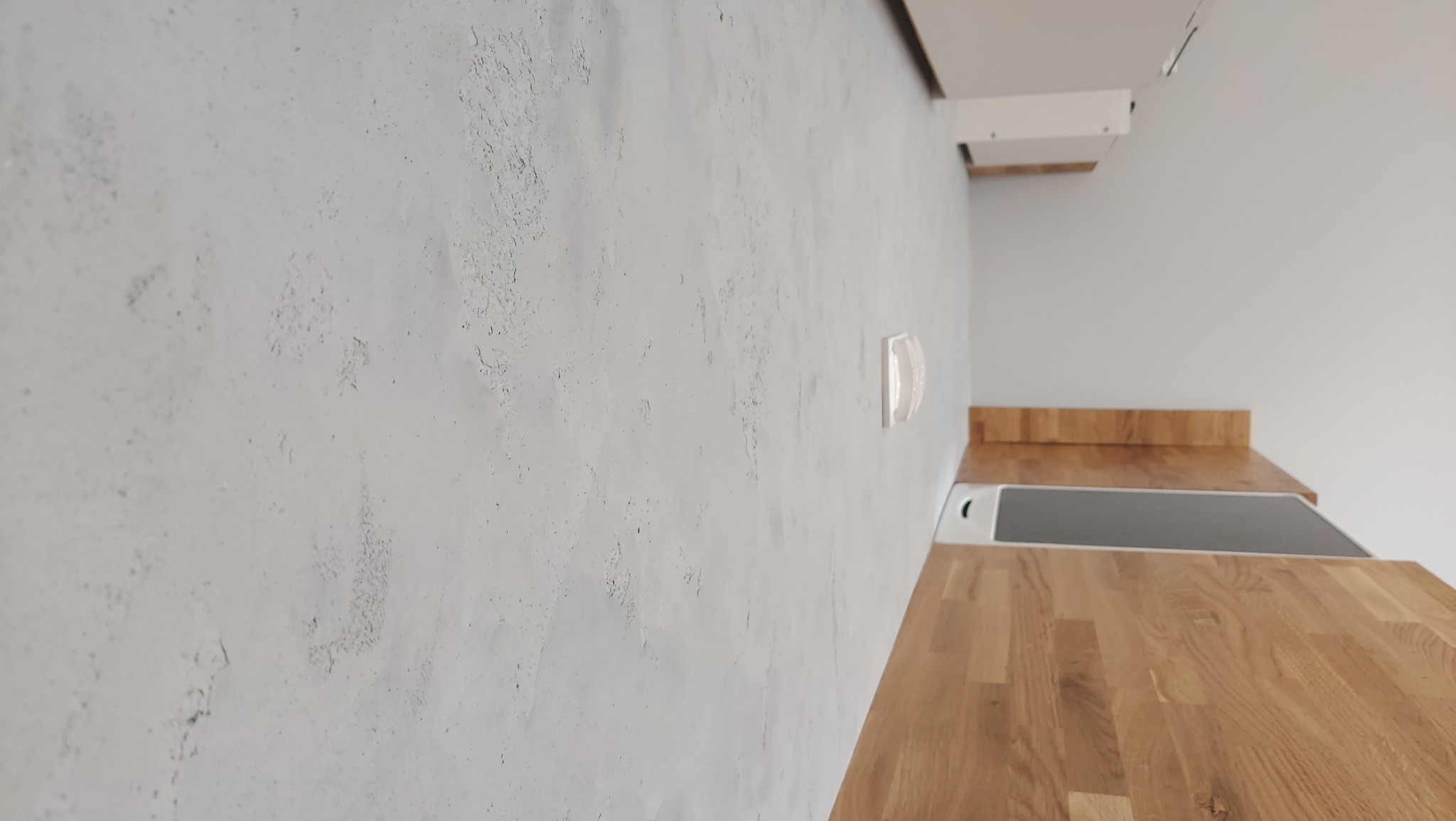 Surowa, szara ściana z widoczną fakturą betonu sąsiaduje z blatem kuchennym z jasnego drewna; widoczne elementy kuchenki i okap.