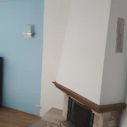 Pomieszczenie po remoncie z dwukolorową ścianą (błękit i biel), kominkiem z kamienia i drewnianym obramowaniem, drewnianą podłogą i nowoczesnym kinkietem.