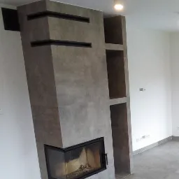 Nowoczesny kominek obłożony szarą płytką, z czarnymi akcentami i otwartą wnęką na drewno, osadzony na podłodze wyłożonej szarymi płytkami w minimalistycznym wnętrzu.