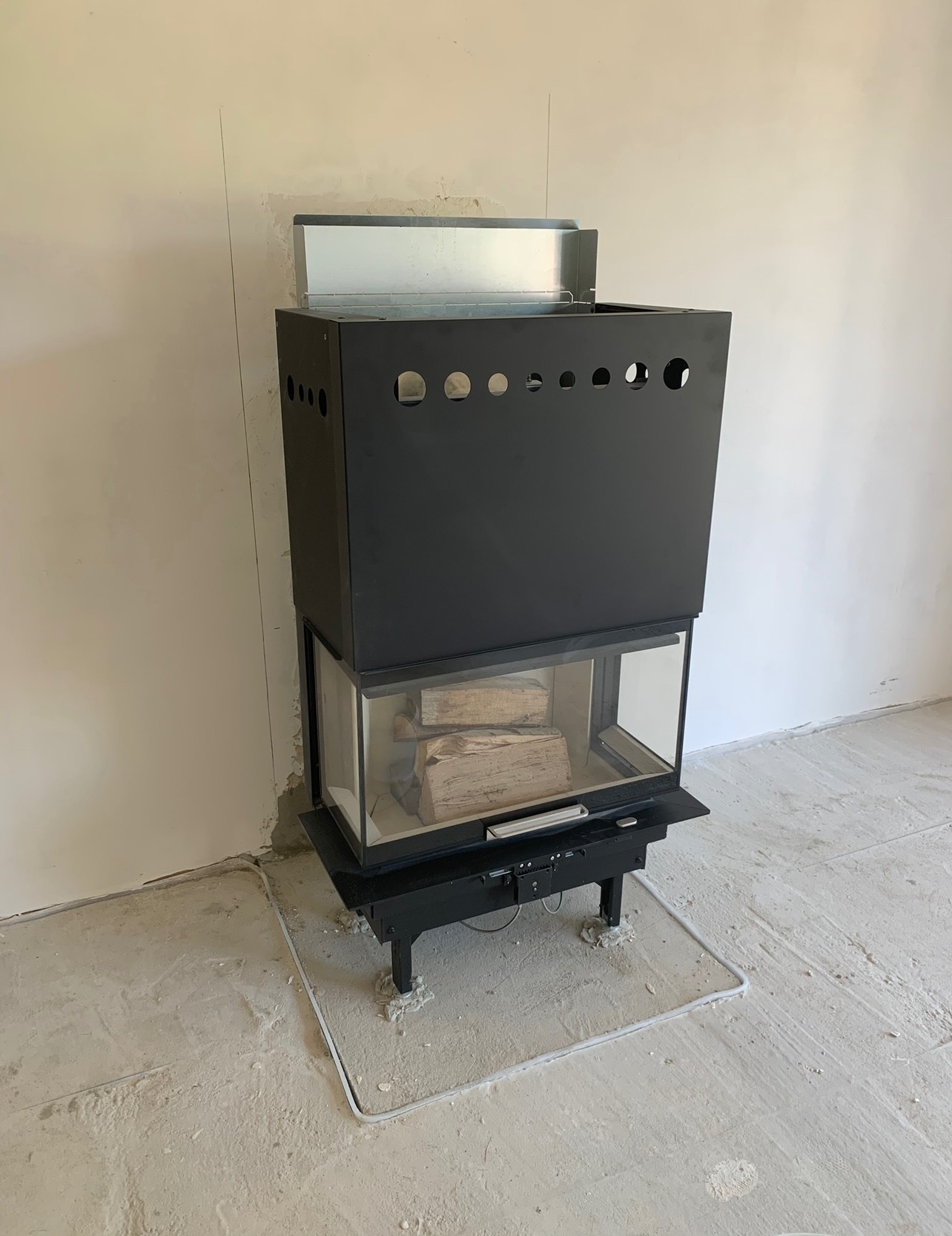 Czarny, nowoczesny kominek z szybą i drewnem w surowym wnętrzu, widoczne ślady prac wykończeniowych na ścianach i podłodze.