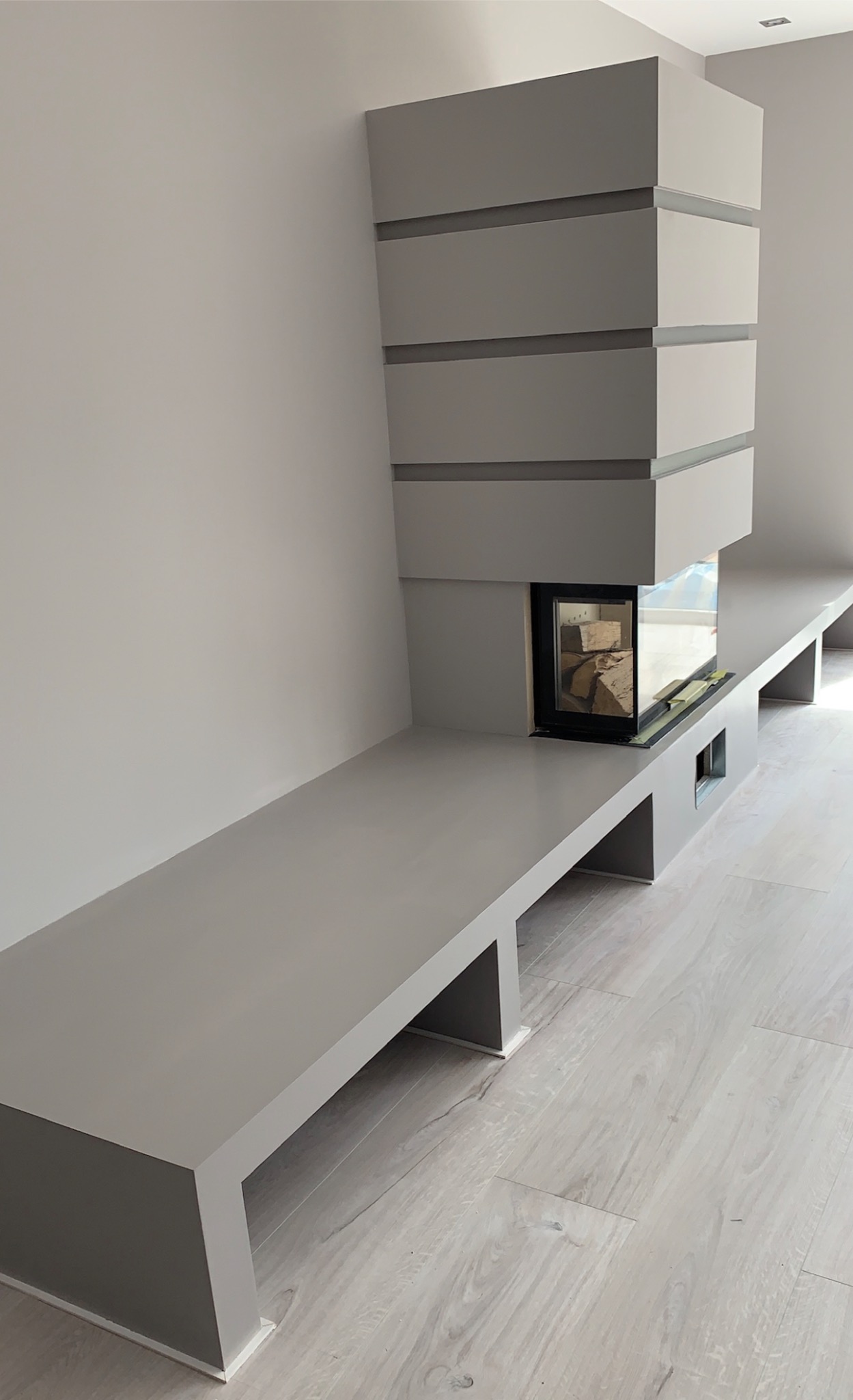 Nowoczesny, minimalistyczny kominek w zabudowie z szarej płyty MDF, z paleniskiem wypełnionym drewnem i długim, niskim blatem po bokach, na tle jasnoszarej ściany i podłogi imitującej drewno.