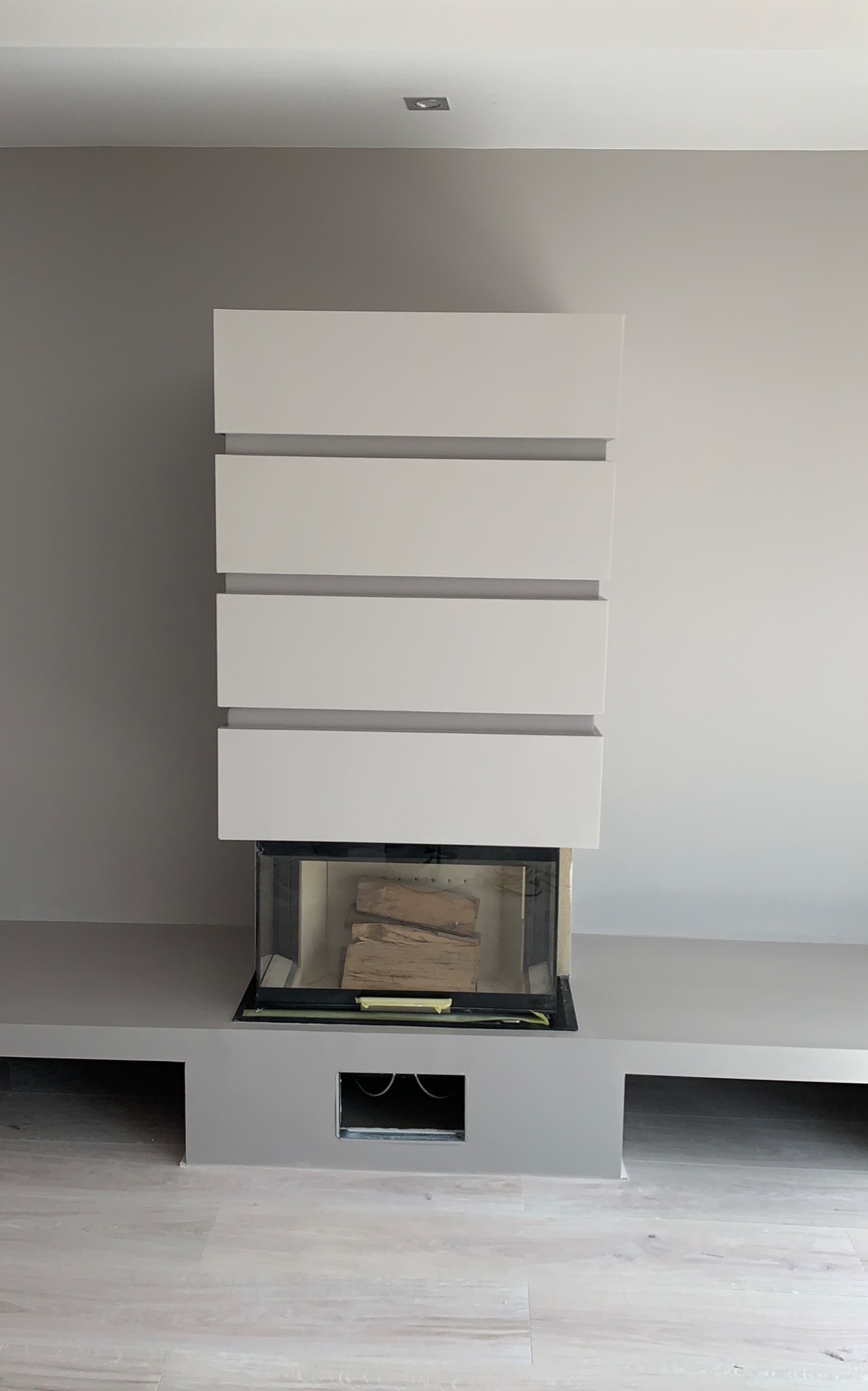 Nowoczesny kominek z wkładem i dekoracyjnym obudowaniem z poziomych, białych paneli, ustawiony na szarej podstawie z wnękami, na tle szarej ściany i jasnej podłogi.