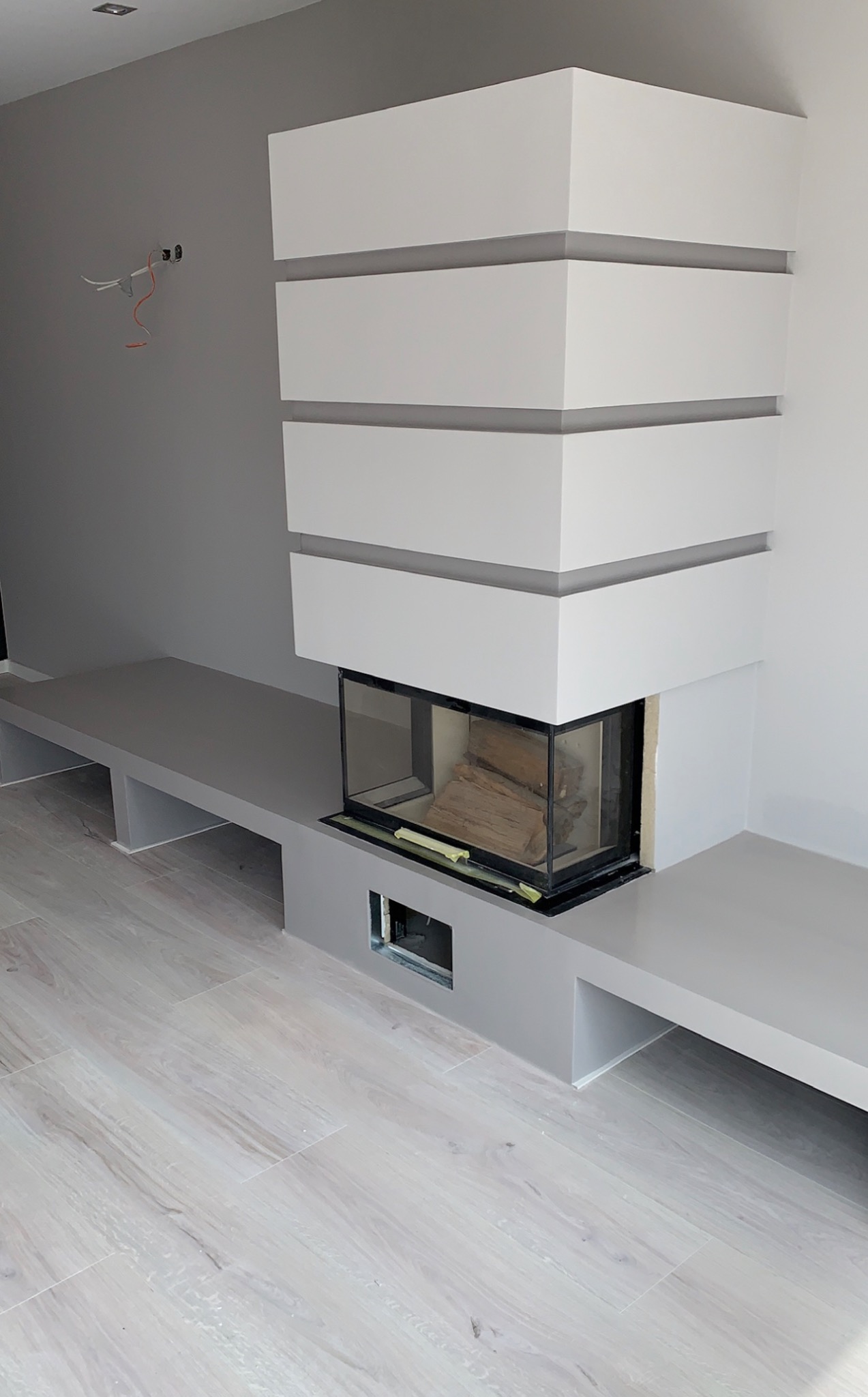 Nowoczesny kominek z obudową w odcieniach szarości i bieli, z widocznymi przewodami elektrycznymi na ścianie, sugerującymi etap prac wykończeniowych.