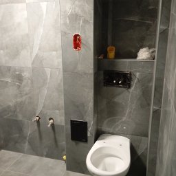 Michał Majcher - Niedokończona łazienka z szarymi płytkami imitującymi marmur, wisząca toaleta bez deski, puszka elektryczna wystająca ze ściany i niezamaskowane rury doprowadzające wodę.