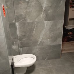 Michał Majcher - Świeżo wyremontowana łazienka z szarymi płytkami imitującymi marmur, podwieszana toaleta bez deski, widoczne kable wystające ze ściany.