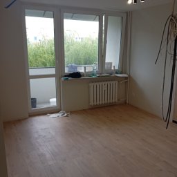 Michał Majcher - Pusty pokój po położeniu paneli podłogowych, widoczne okno z widokiem na zieleń, grzejnik pod oknem i lampy sufitowe.