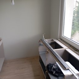 Michał Majcher - Montaż szafek kuchennych pod oknem, widoczne szyny montażowe i przewody elektryczne, surowy stan wykończenia.