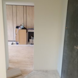 Michał Majcher - Widok przez drzwi na remontowane pomieszczenie z drewnianą podłogą i marmurową posadzką przy wejściu, widoczne narzędzia i materiały budowlane.