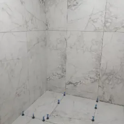 Białe płytki imitujące marmur ułożone w rogu łazienki, z niebieskimi klipsami poziomującymi, widoczne fugi.