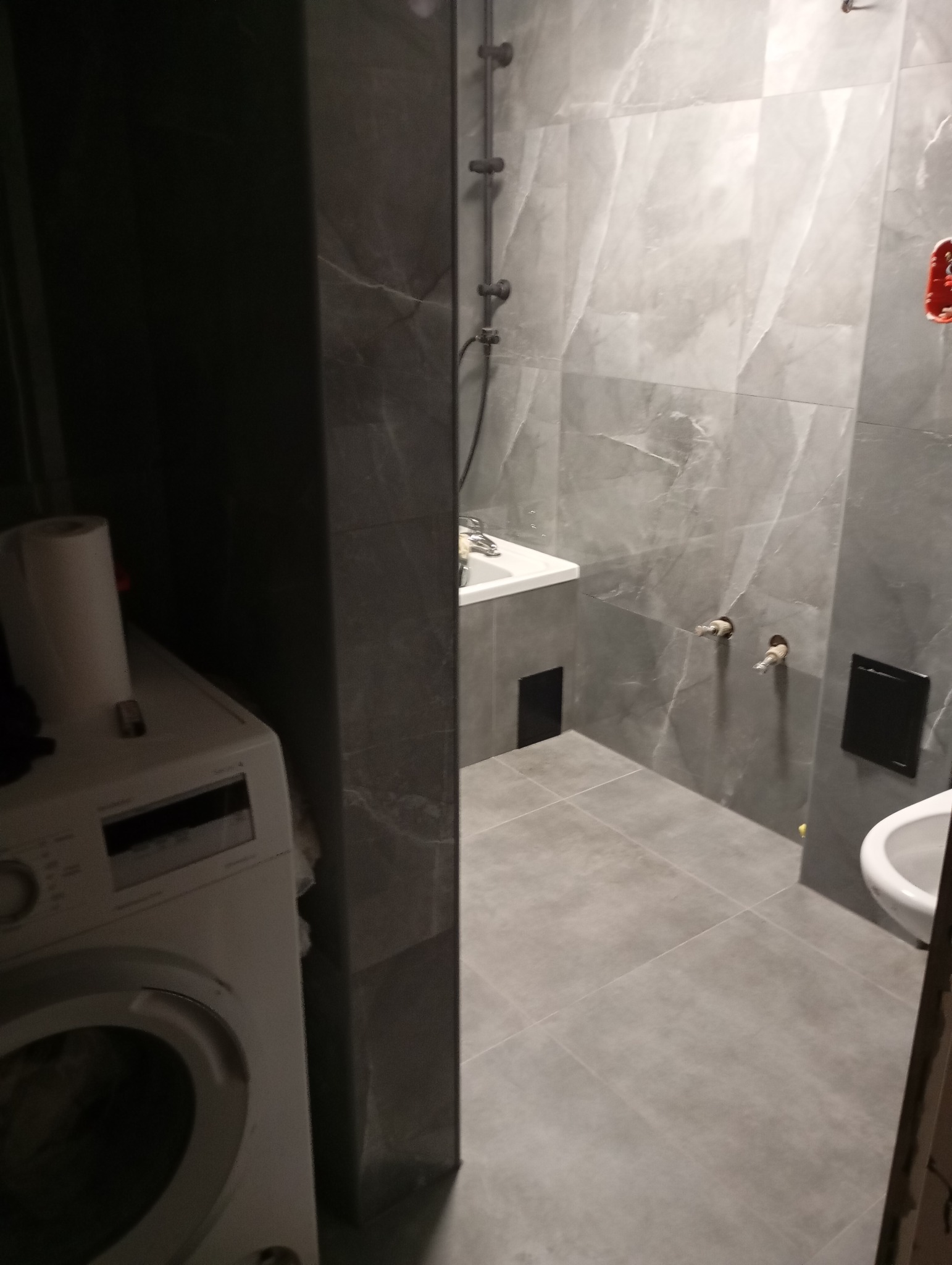 Szara łazienka w trakcie wykańczania, widoczna pralka, umywalka, toaleta i płytki imitujące marmur, z wystającymi rurami w ścianie.